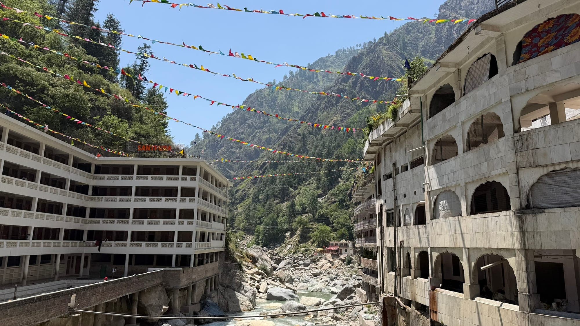 Lines of small coloured flags strung high across a narrow river gorge between a white multi-storey building with a Santpura sign on the left and an arched, weathered building on the right, with green mountains and a rocky stream below