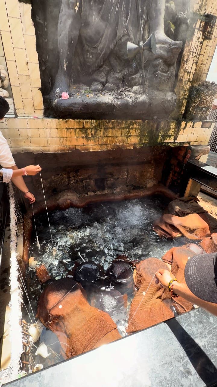 A stone tank of bubbling, steaming spring water below a dark stone statue, with strings and cloth bundles lowered into the water by hands reaching in from both sides