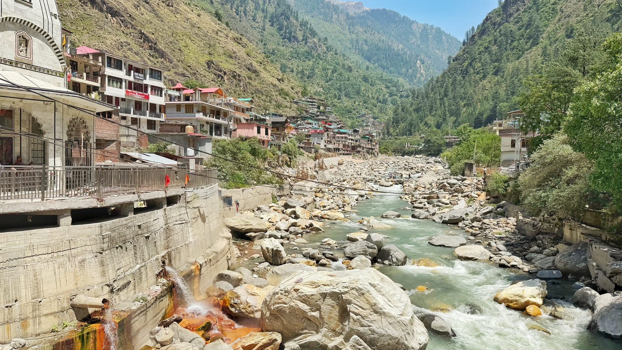 The town of Manikaran climbing the left bank of the Parvati river, its guesthouses stacked on the hillside above a temple with a railing, with the river tumbling over boulders in the centre and orange-stained water running down the retaining wall at the bottom left