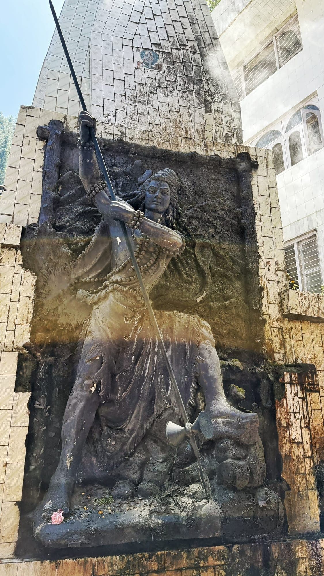 A dark relief statue of Shiva with matted hair, rudraksha beads and a cobra, holding a long trident with a damaru, set against a stone wall in front of a tiled temple spire, with a pink flower and coins left at the base