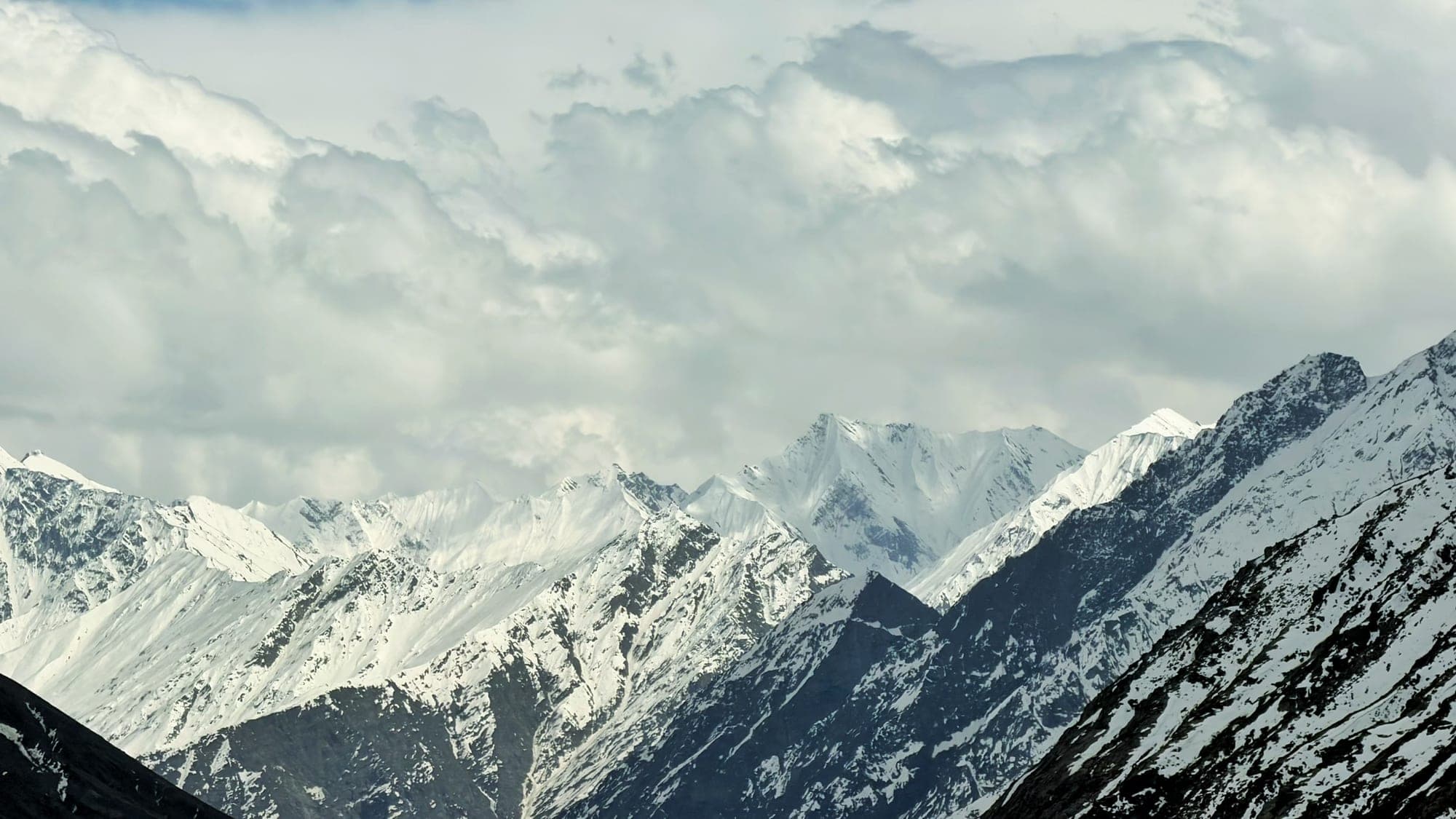 A ridge of sharp white snow peaks under heavy grey cloud, with a dark, snow-streaked mountain in the foreground at the right