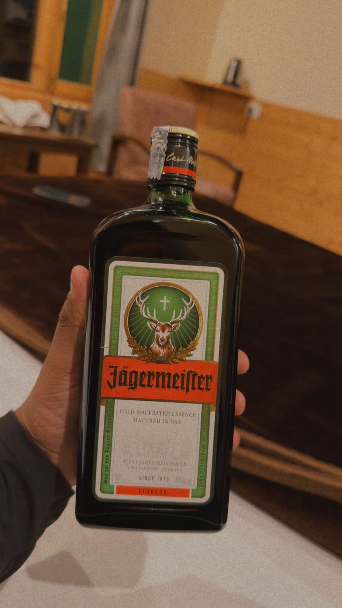 A hand holding up a large bottle of Jägermeister, label facing the camera, in a wood-panelled room with a pink chair and tables blurred behind