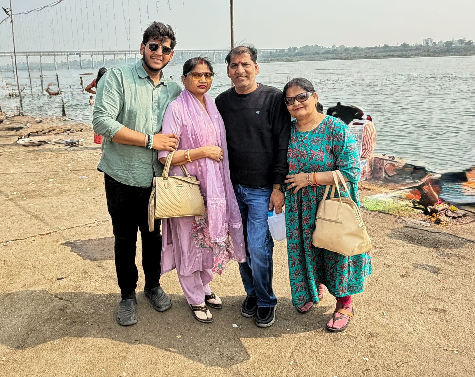 Four people standing together on a sandy riverbank, with a wide river and a long bridge behind them