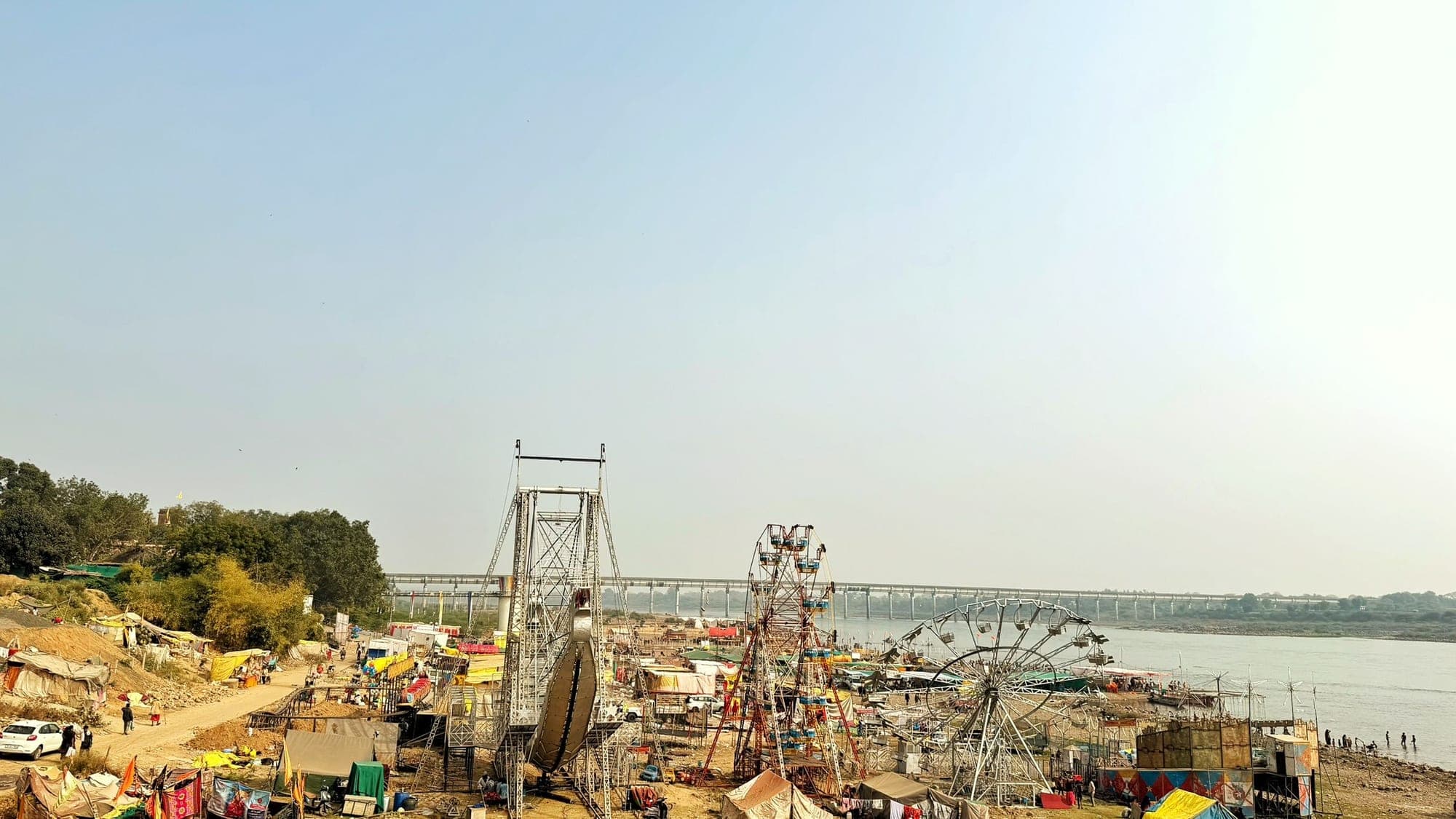 A wide river with a long bridge in the distance, and a small fair on the bank with a Ferris wheel, stalls and tents