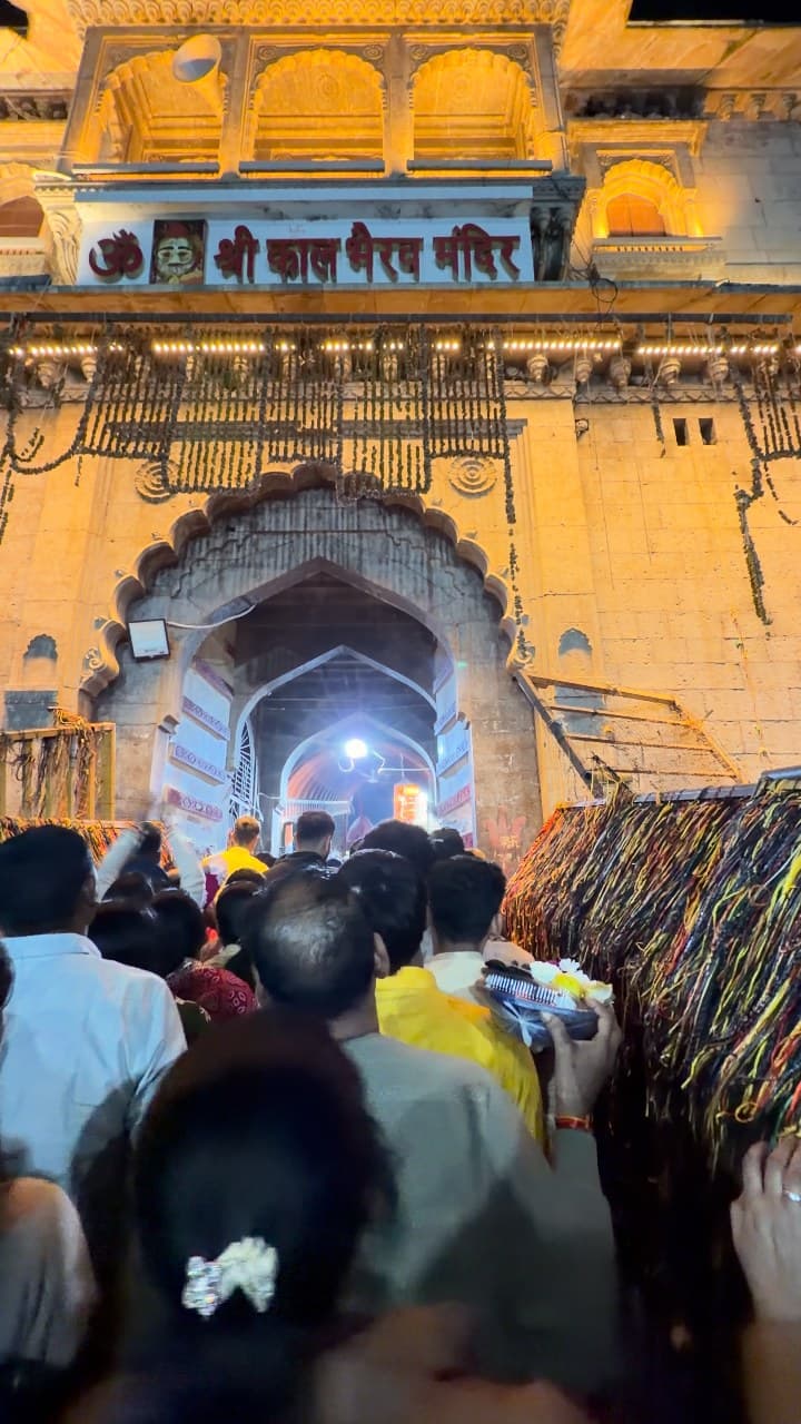 A tight crowd moving towards the lit gate of the Shri Kaal Bhairav Mandir at night, with coloured threads hanging at the right