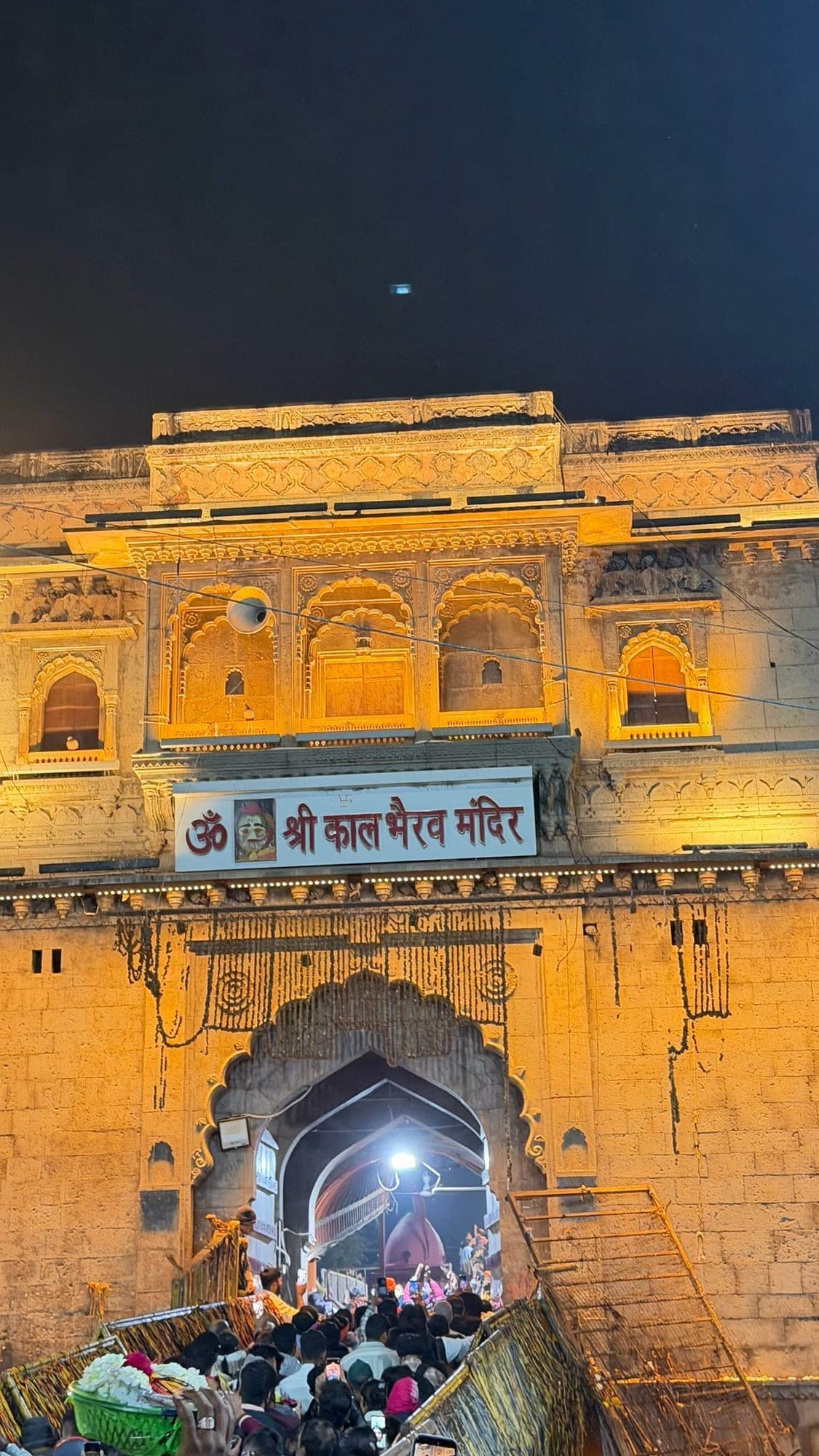 The yellow sandstone gate of the Shri Kaal Bhairav Mandir lit up at night, with a crowd climbing the steps to its arched doorway