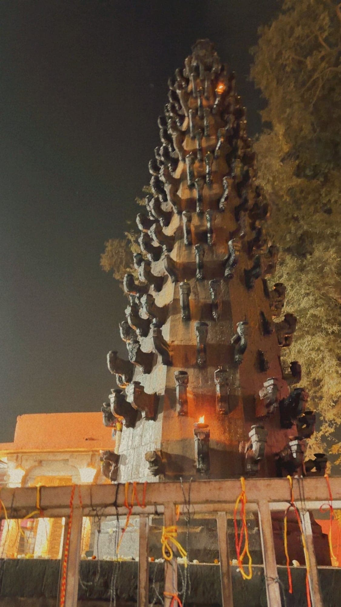 A tall, tiered stone lamp tower at night, its rows of small niches holding lit diyas, in front of an orange-lit building