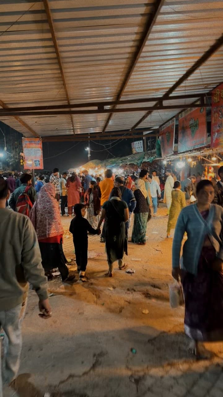 A crowded lane at night under a metal roof, with people walking in both directions and market lights ahead