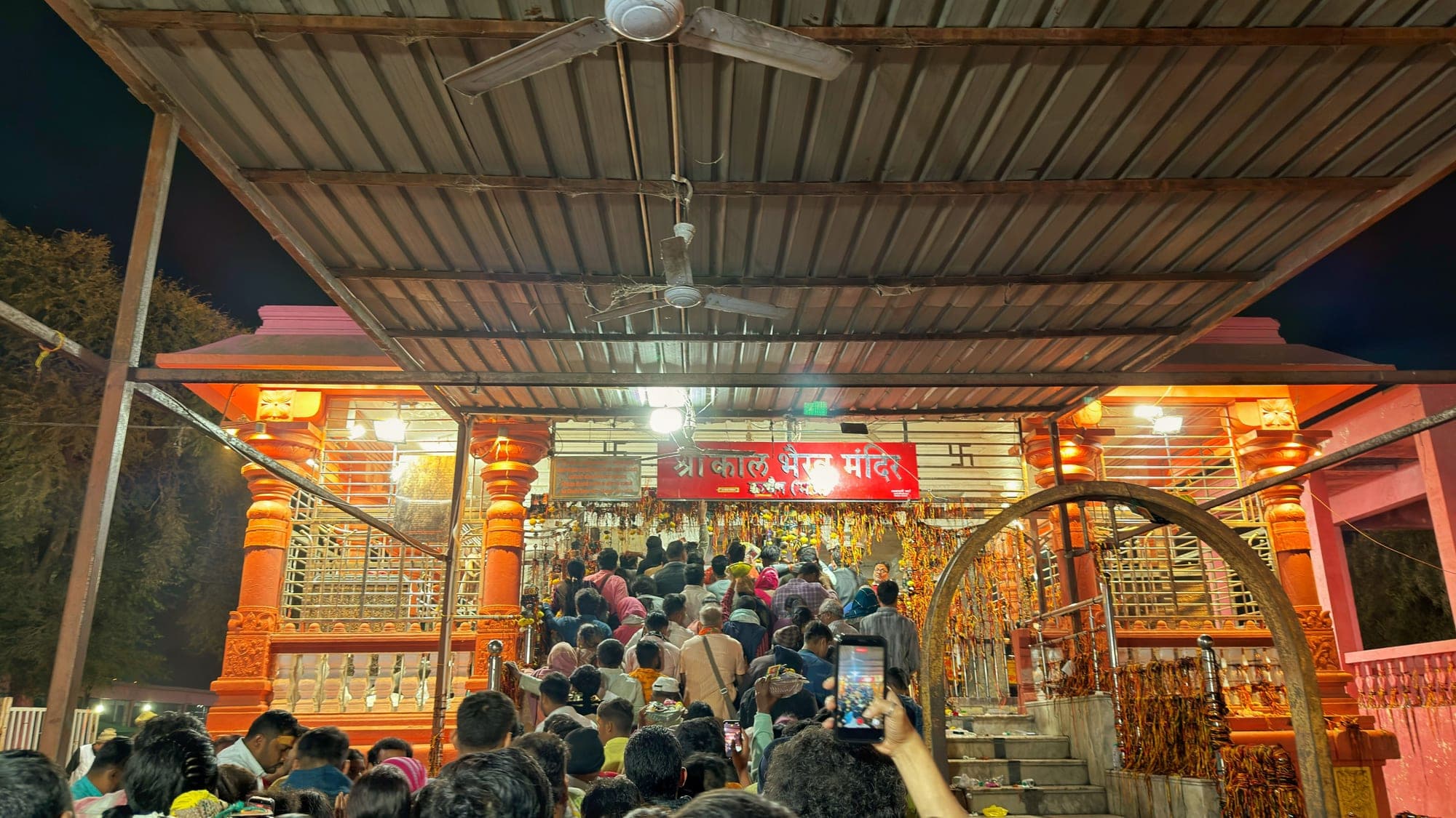 A packed queue of people waiting at the orange-painted entrance of the temple's inner shrine at night, under a tin roof, with a phone raised at the right