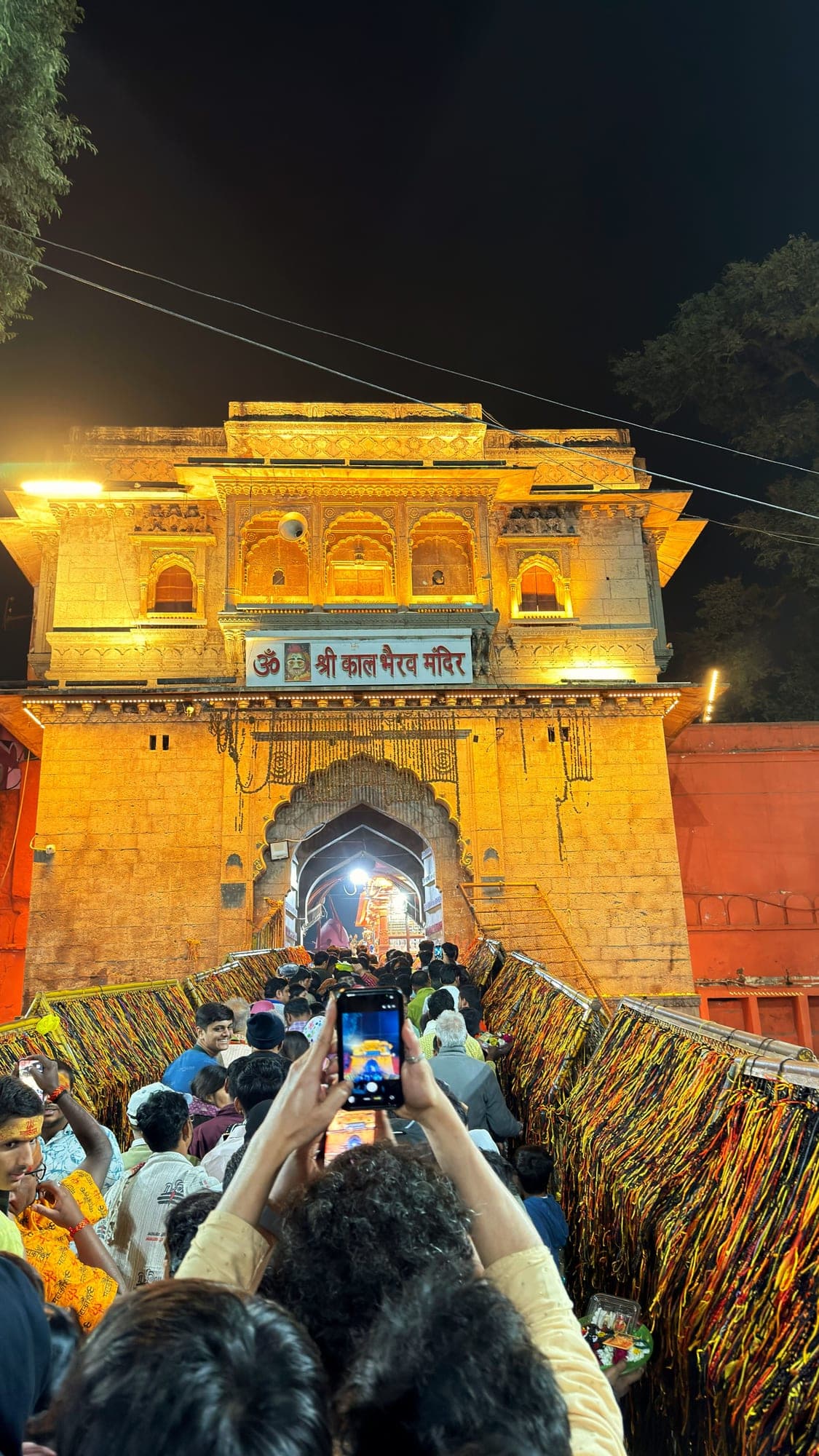 A crowd climbing the steps to the Kaal Bhairav gate at night, seen from behind, with a phone held up in the middle and threads hanging on the railings on both sides