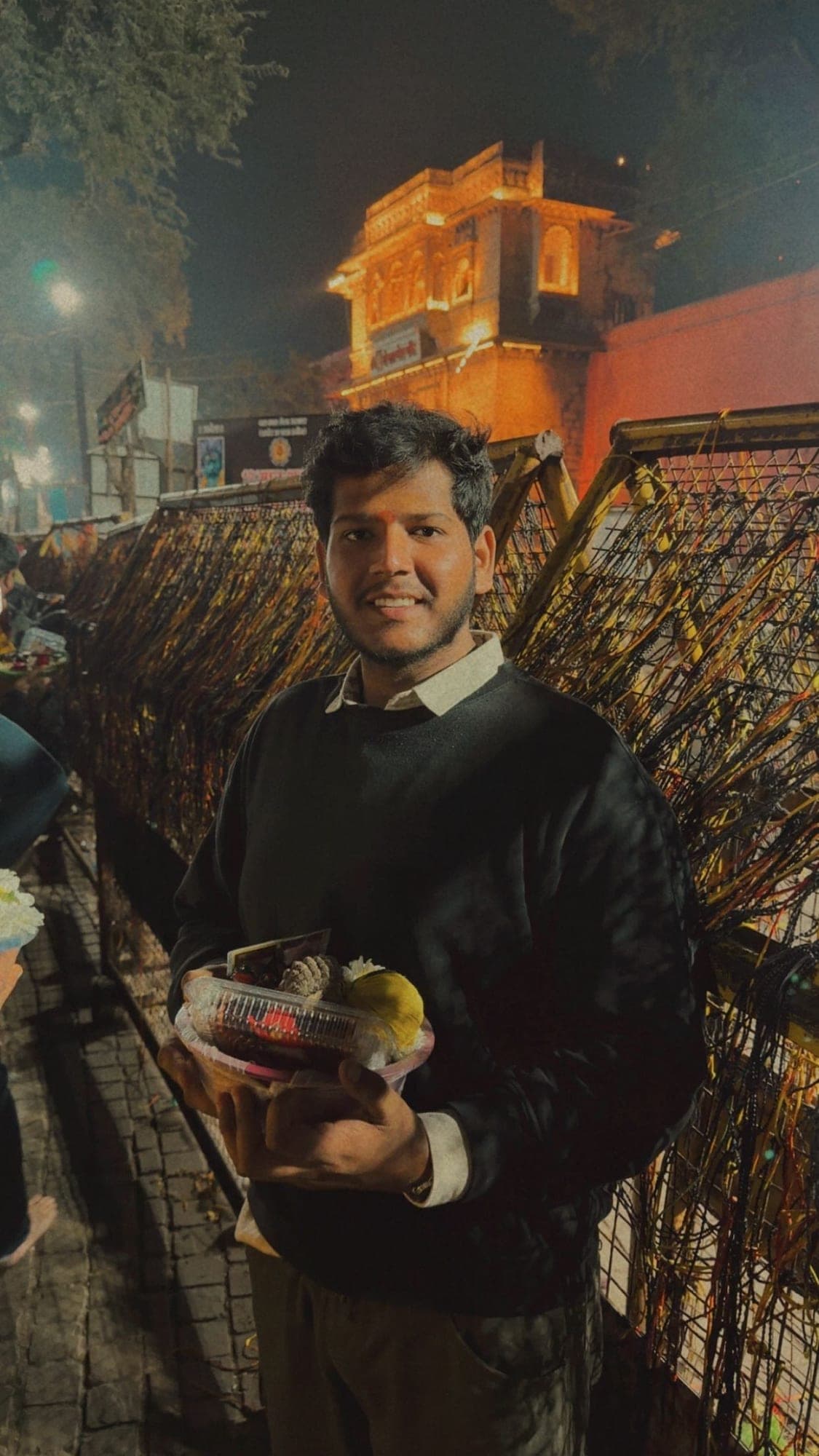 A young man in a dark sweater holding a basket of offerings beside a railing woven with colourful threads, with the lit temple gate behind him