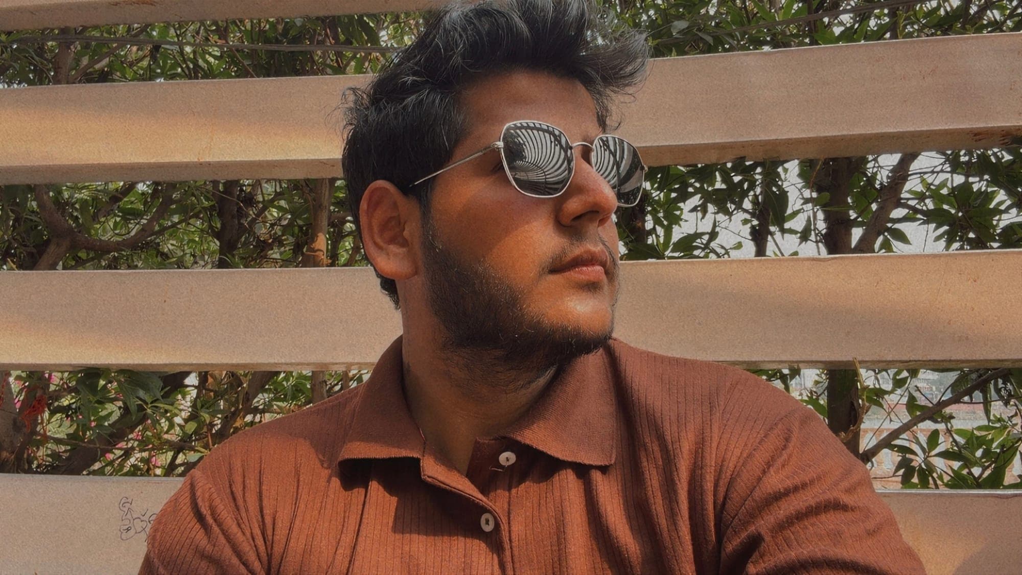 A close portrait of a young man in sunglasses and a brown ribbed polo shirt, looking away to the right, in front of wooden slats and green leaves, with a curved slatted ceiling reflected in the lenses