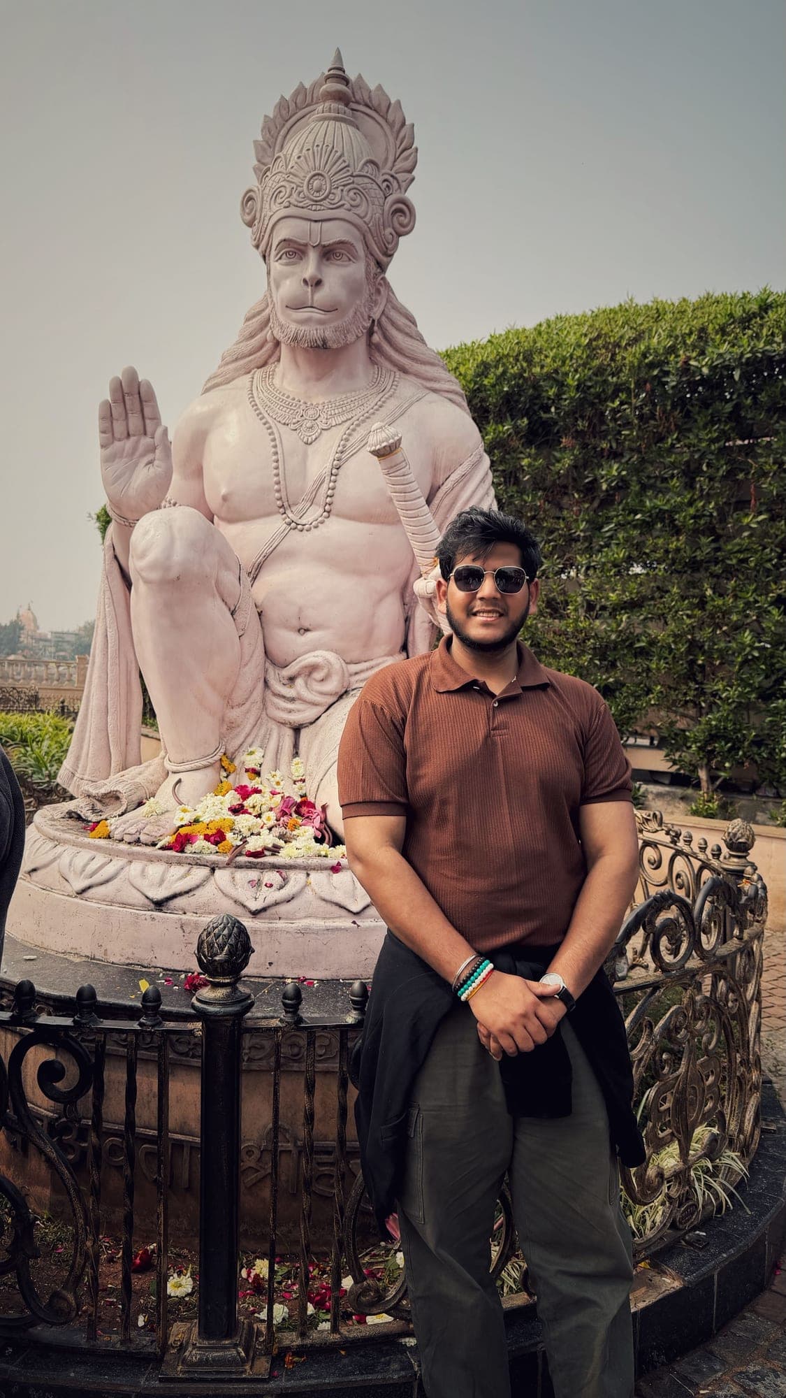 A young man in sunglasses and a brown shirt standing in front of a large pale statue of Hanuman seated with a raised hand, with flowers at its base and an iron railing around it