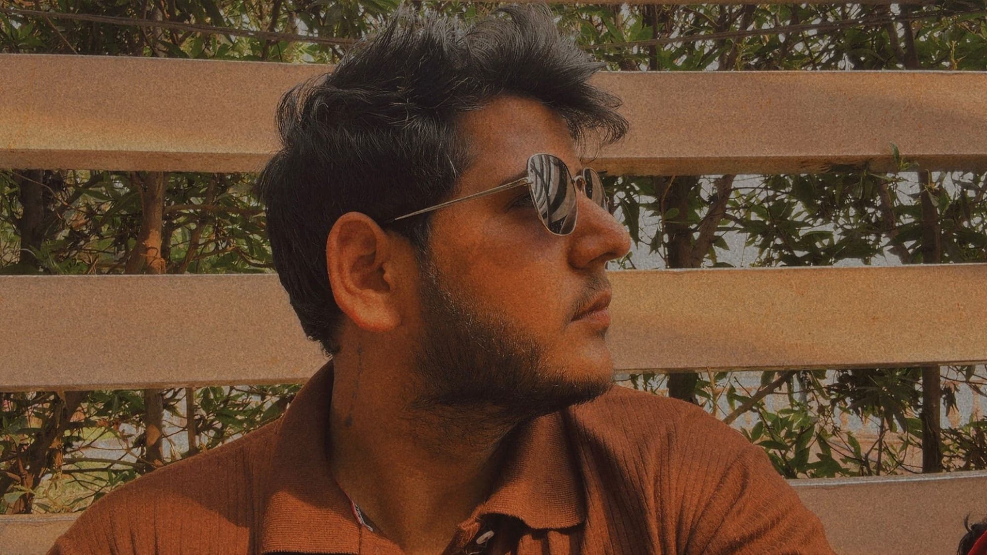 A young man in sunglasses and a brown shirt looking away to the side, in front of a curved wooden slatted screen