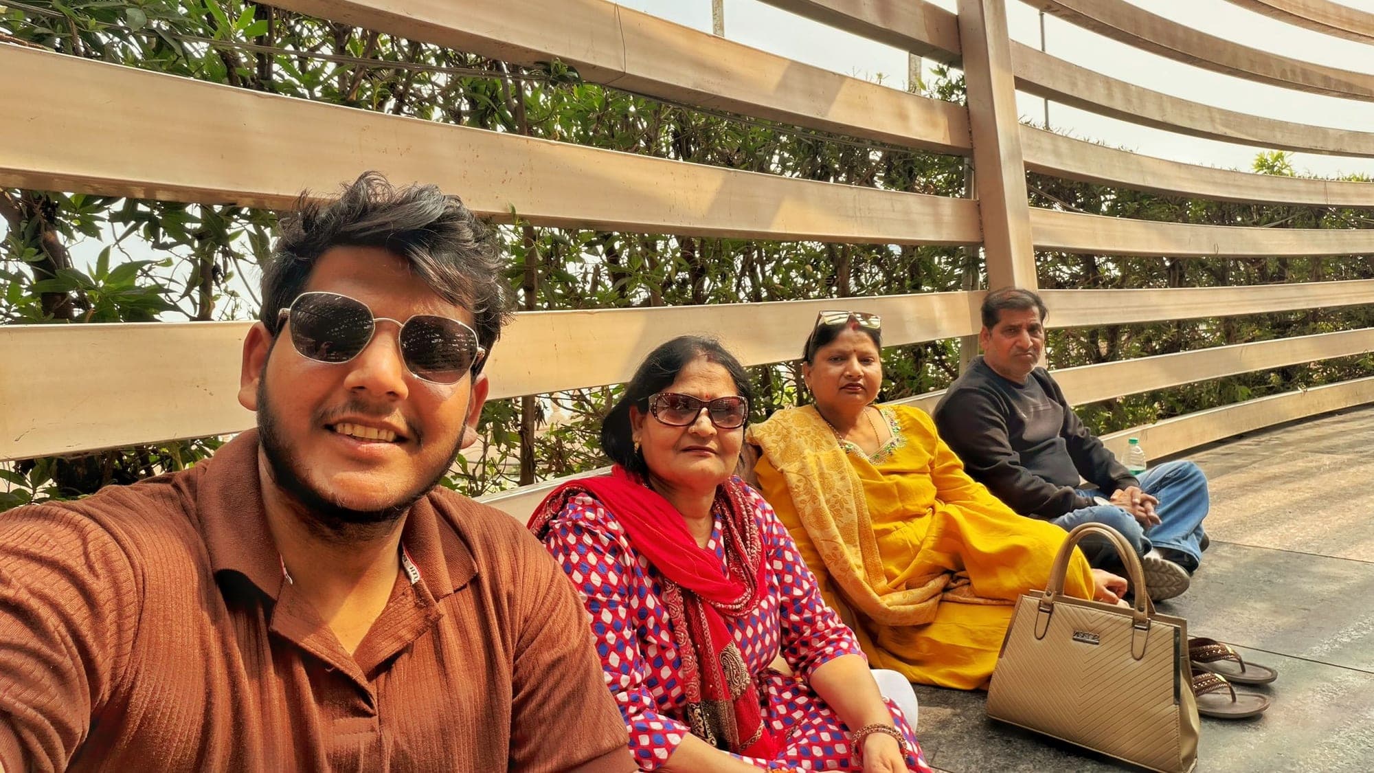 Four people sitting on a curved wooden bench in a slatted shelter, one in a brown shirt and sunglasses holding the camera for a selfie