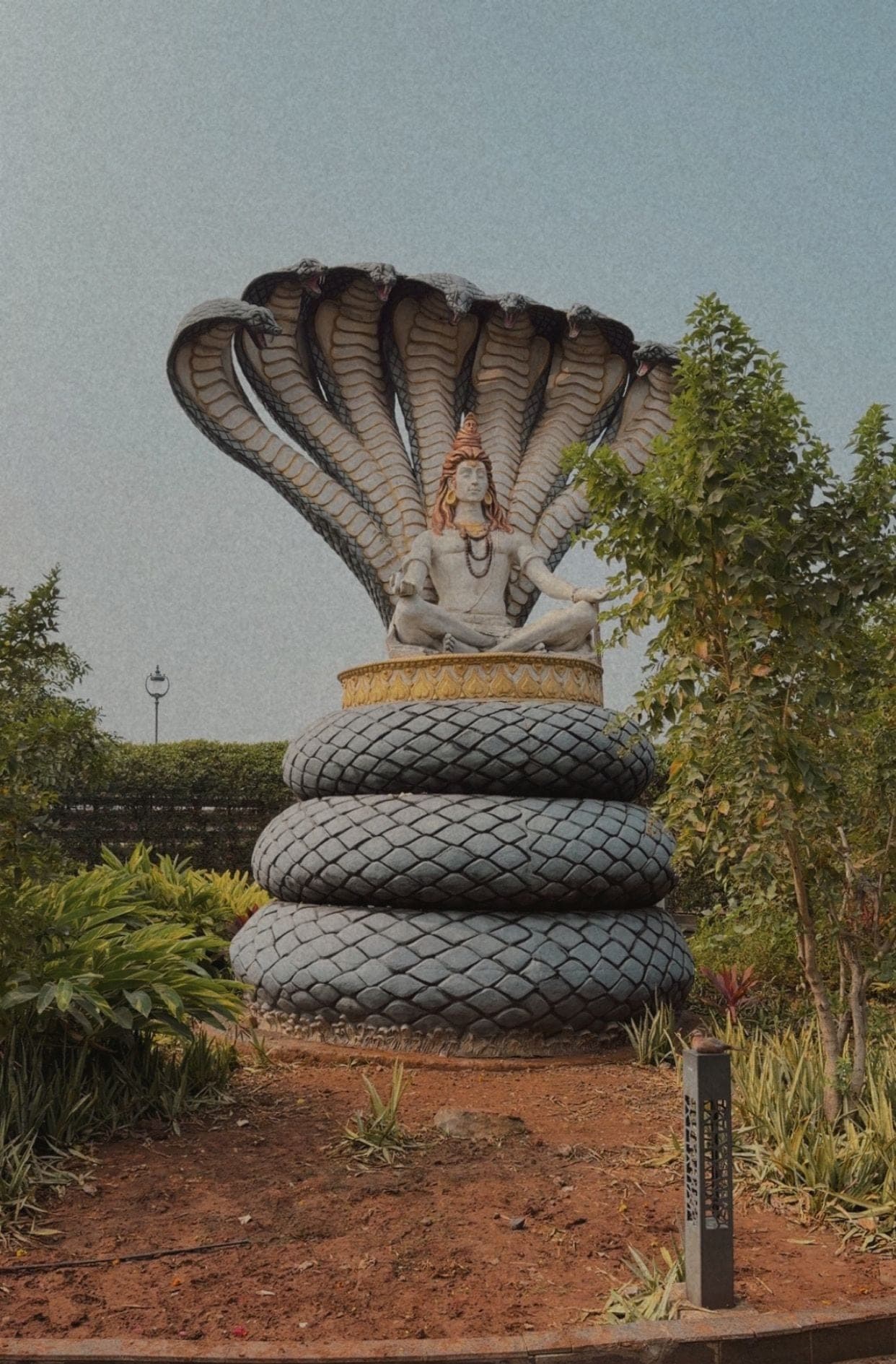 A pale statue of Shiva seated in meditation on three coils of a snake, beneath a spread of many cobra hoods, in a landscaped garden