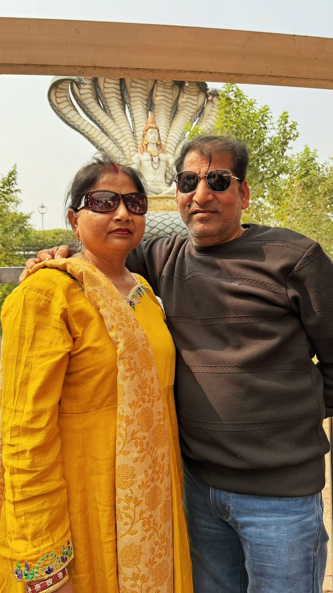 A woman in a yellow outfit and sunglasses and a man in a brown sweater and sunglasses posing together, with the many-hooded serpent statue behind them