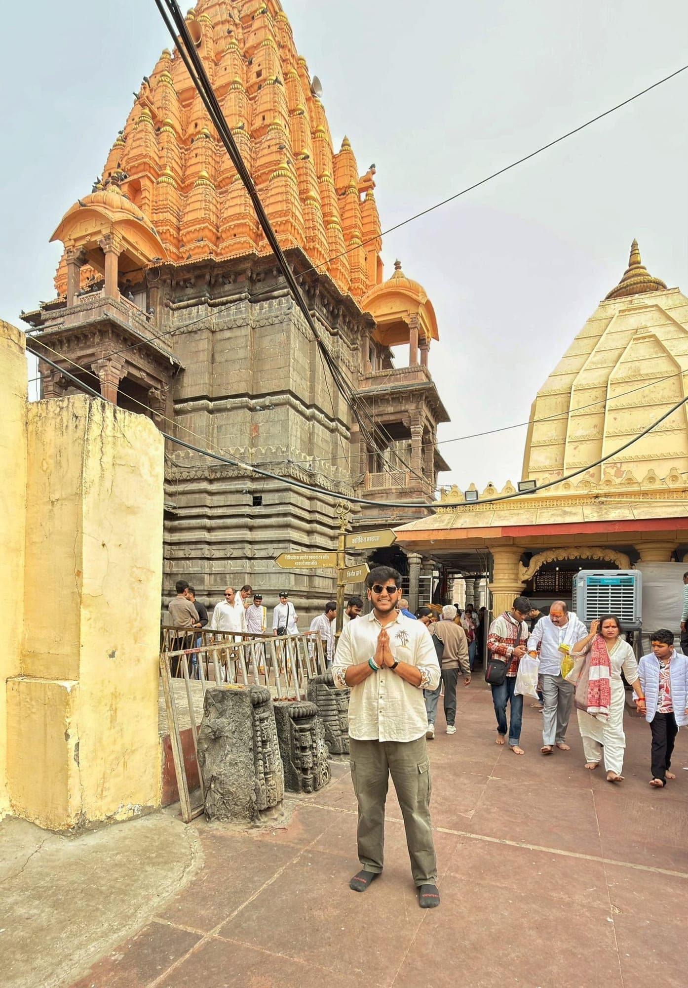 A young man in sunglasses with his palms together in a temple courtyard, with the orange spire behind him and a cream-coloured dome at the right