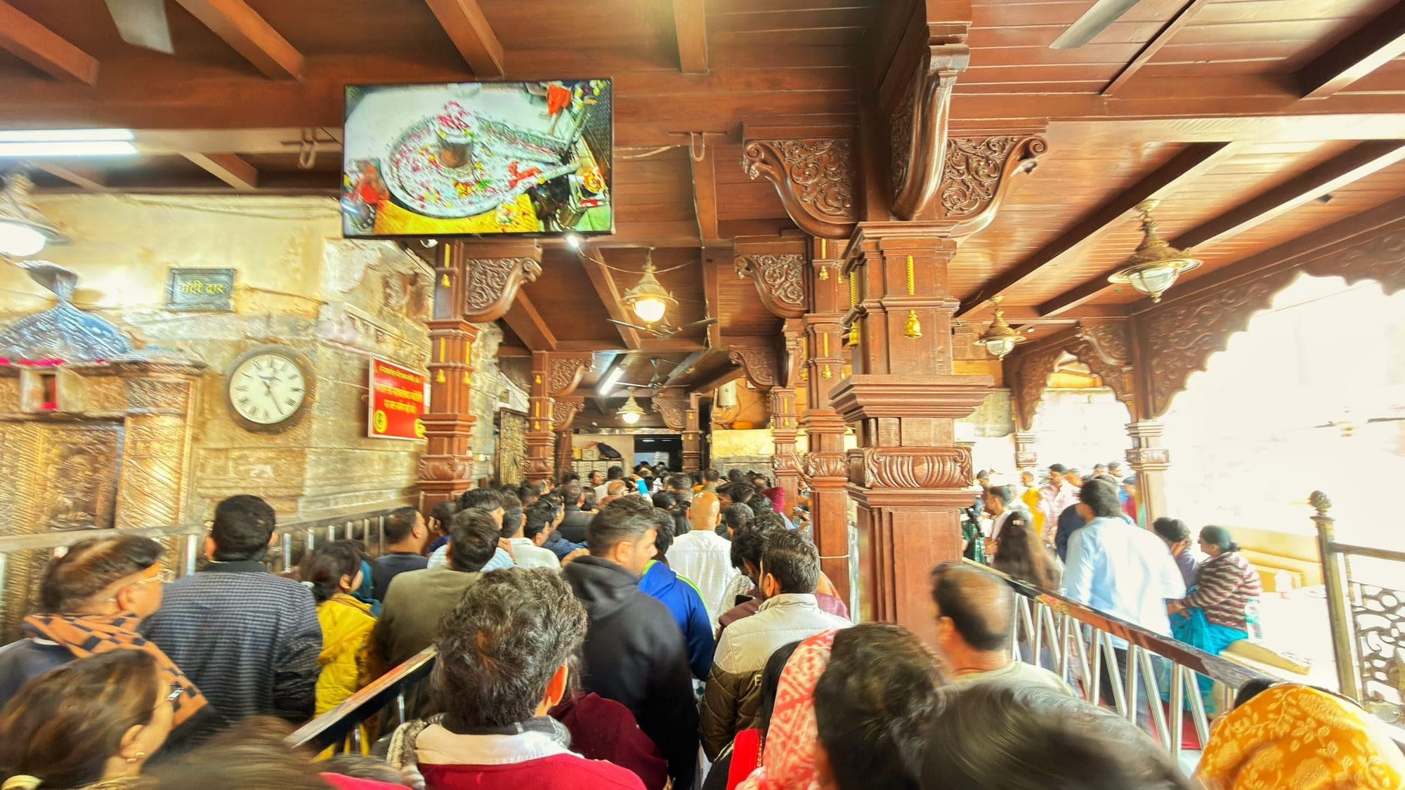 A tightly packed line of people moving through a temple hall with a carved wooden ceiling, a screen showing the lingam above them and a clock on the wall