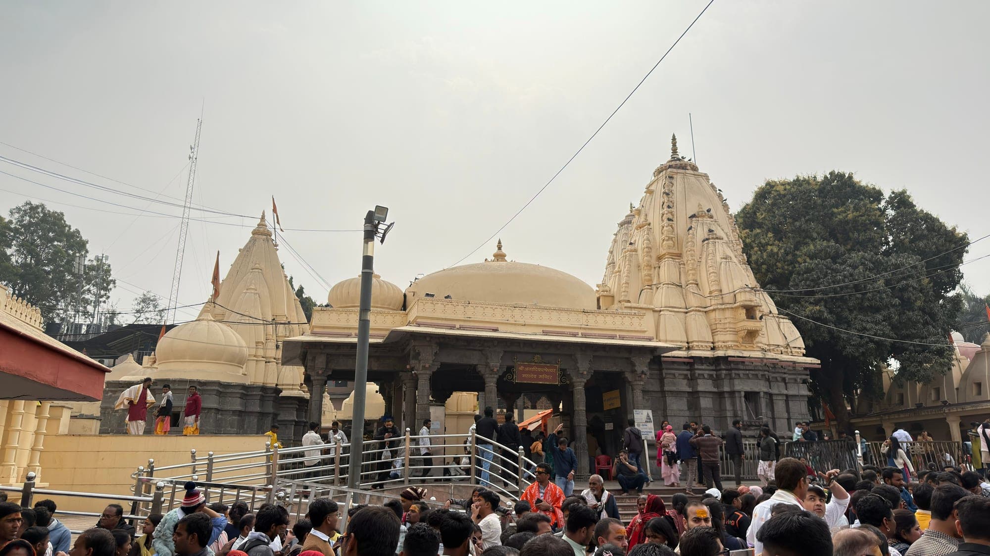 A wide view of the temple complex under a grey sky, with cream-coloured shrines and domes, an open hall and a large crowd in front