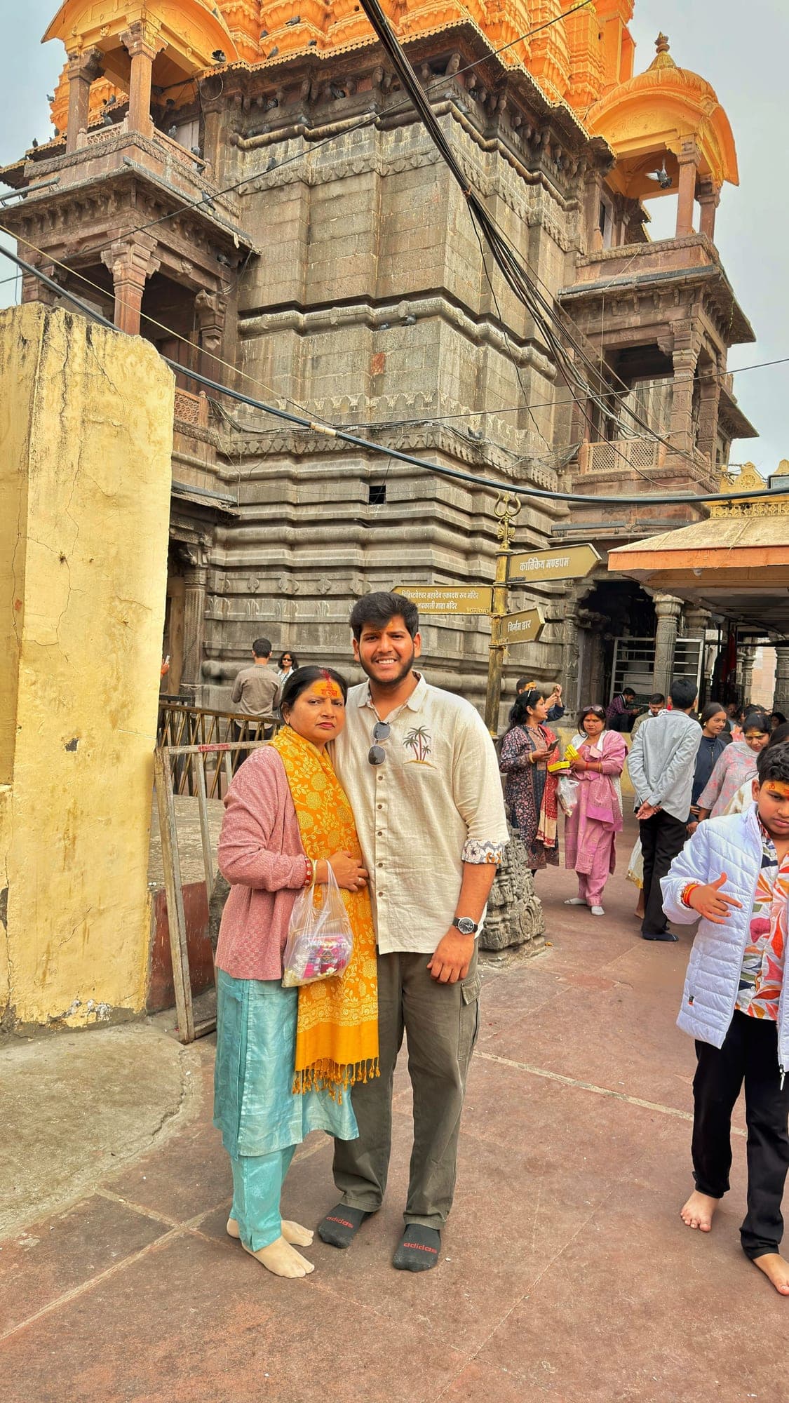 A woman in a pink cardigan with a yellow shawl and a young man in a cream shirt standing together in a courtyard, with the orange spire and dark stone walls of the temple behind them