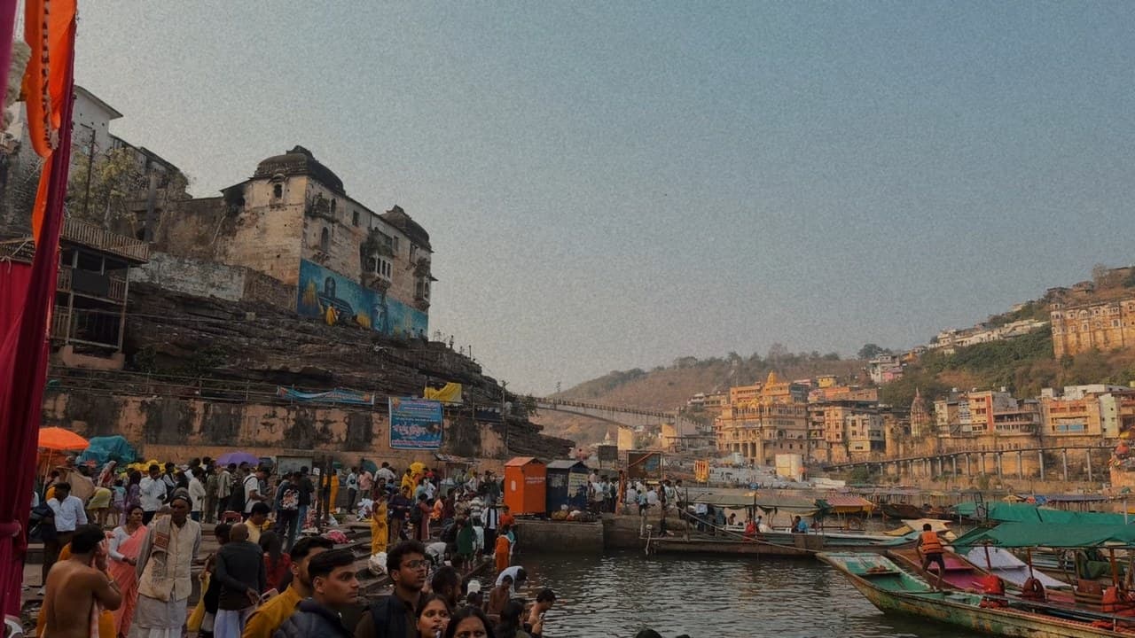 A wide view of the Omkareshwar ghat with a crowd at the water's edge, boats, and old buildings and temples on the hills
