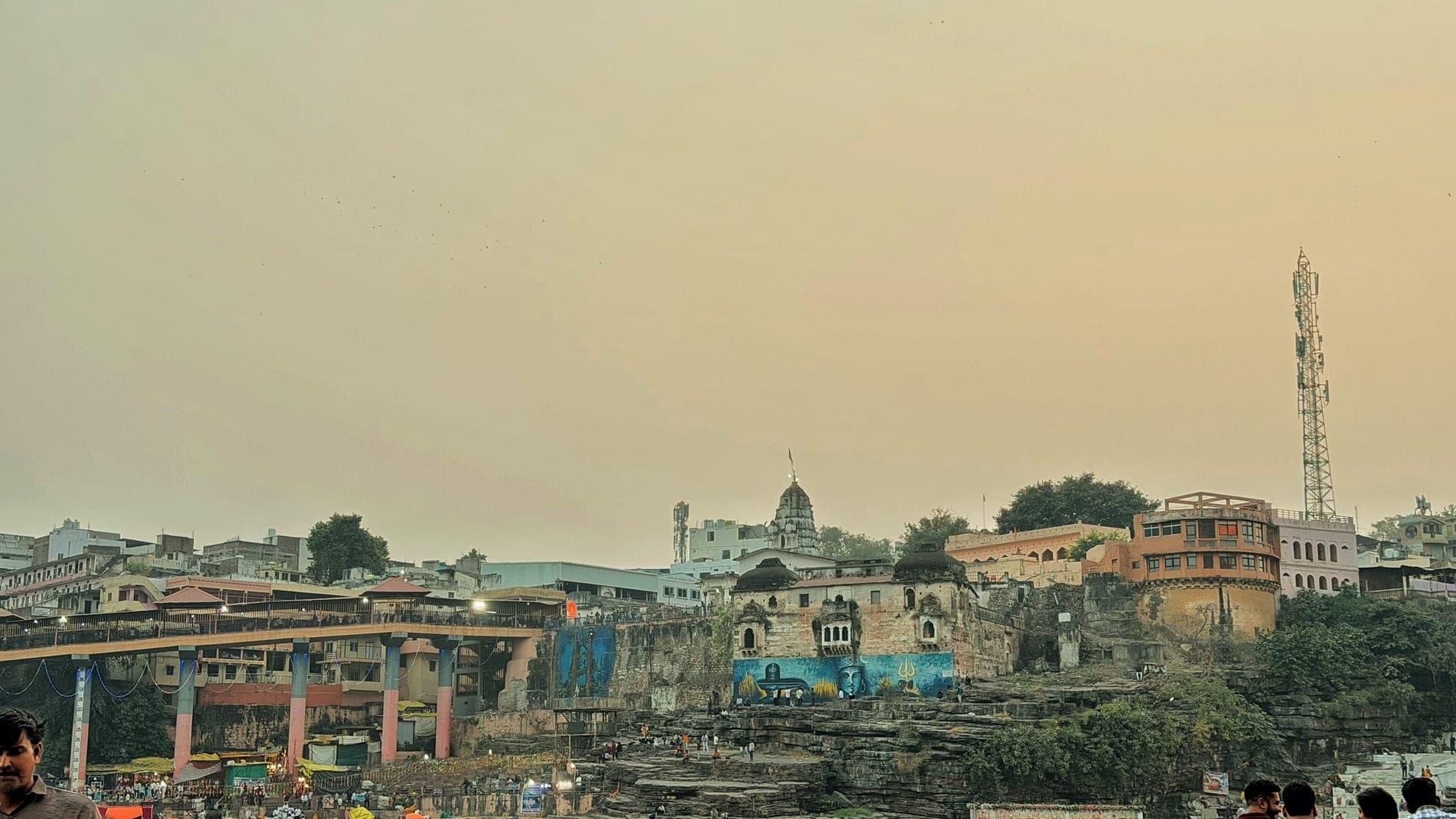 Dusk over Omkareshwar, with a bridge, old stone buildings and a mobile tower on the far bank under a hazy yellow sky