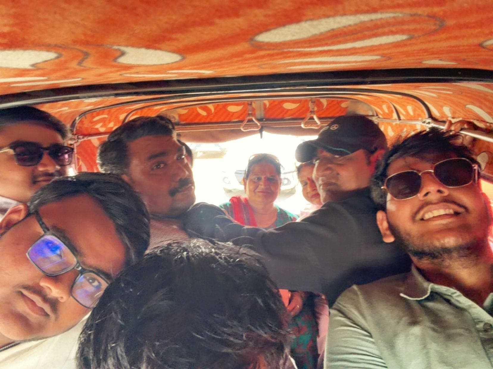 Many people squeezed into the back of an auto-rickshaw under an orange patterned roof, faces close together, one in sunglasses smiling widely at the front