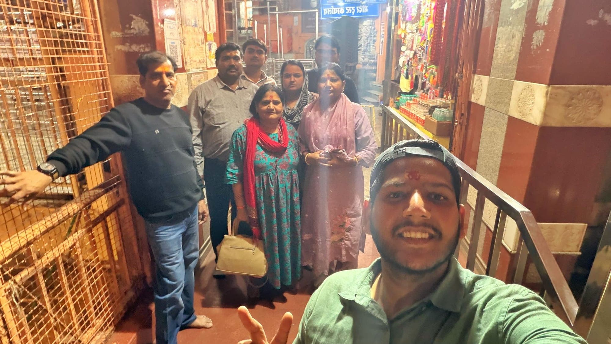 A group of six people standing in a narrow temple lane lined with shops, the young man at the front, in a green shirt with a tilak, holding the camera for a selfie
