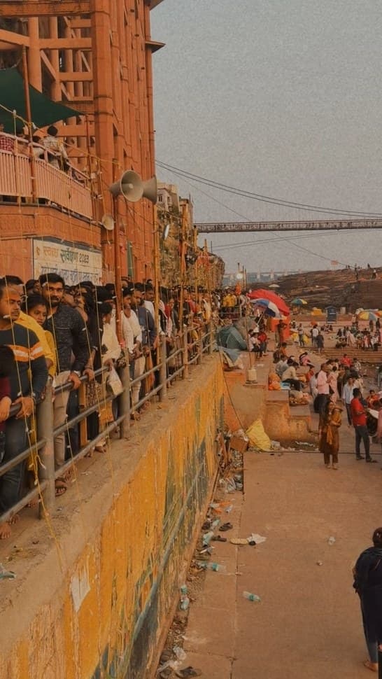 A queue of people standing behind a railing along the ghat, with a suspension bridge in the distance under a dusty sky