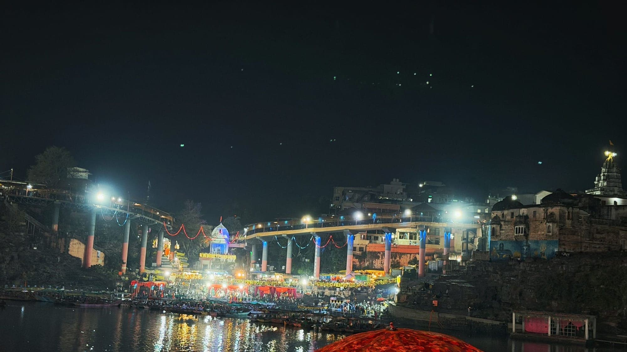 Omkareshwar at night, with brightly lit bridges, a crowd at the water and strings of lights reflected in the dark river