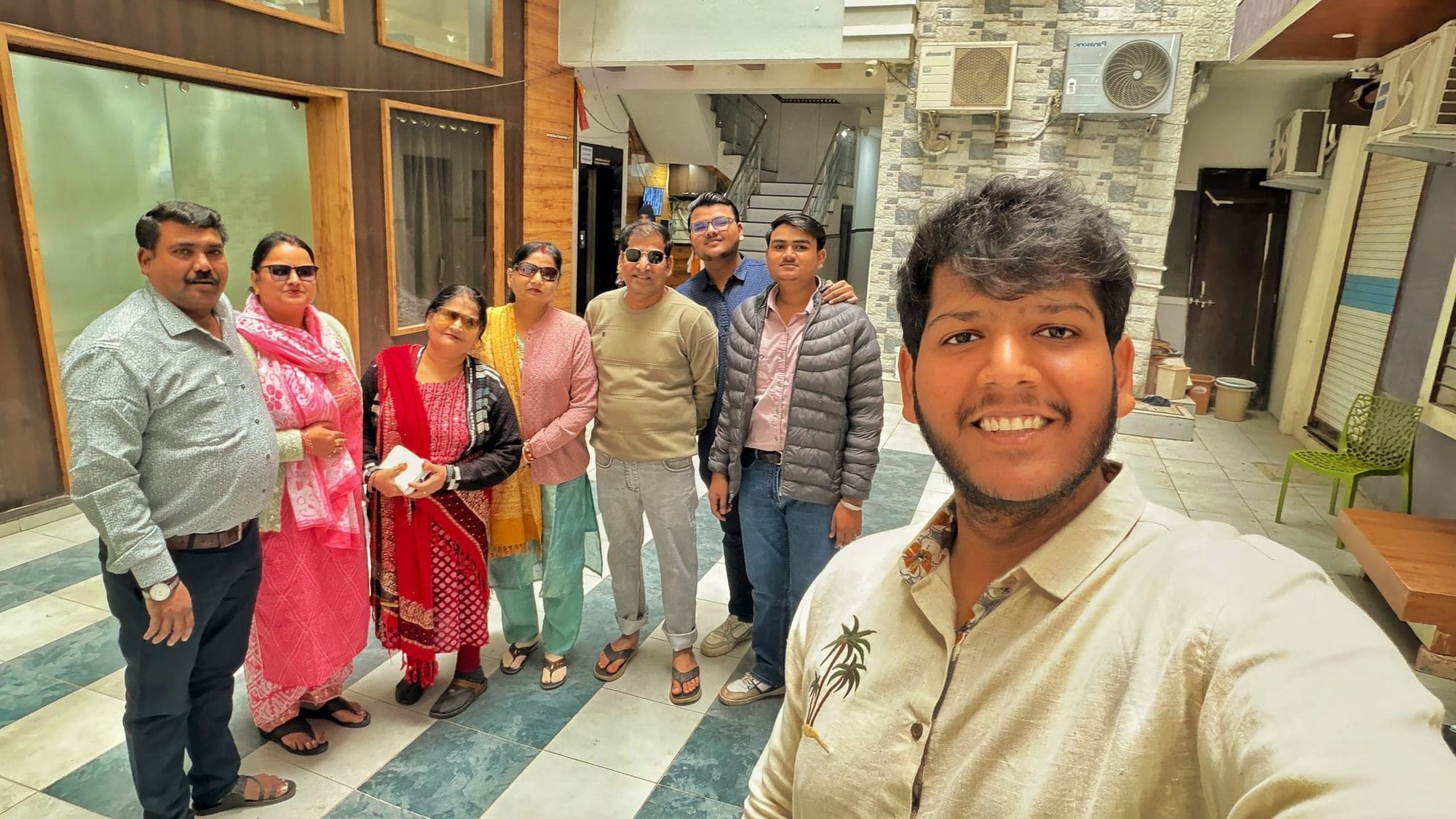 A group of seven people standing close together in a bright courtyard with stairs and balconies behind them, smiling, one of them at the right holding the camera for a selfie