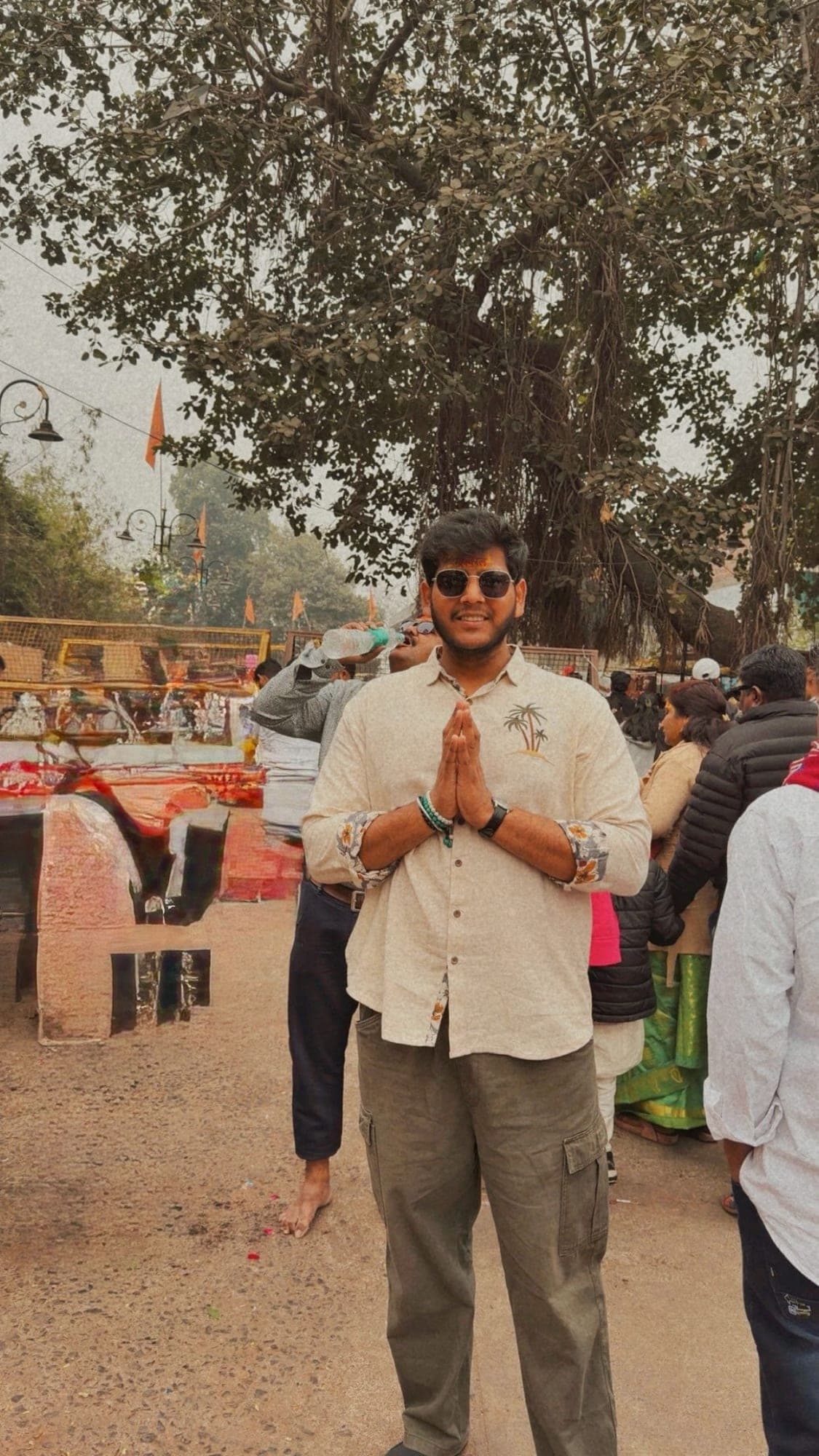 A young man in sunglasses and a cream shirt standing with his palms together in front of a large banyan tree, on a road with barricades and a crowd behind him