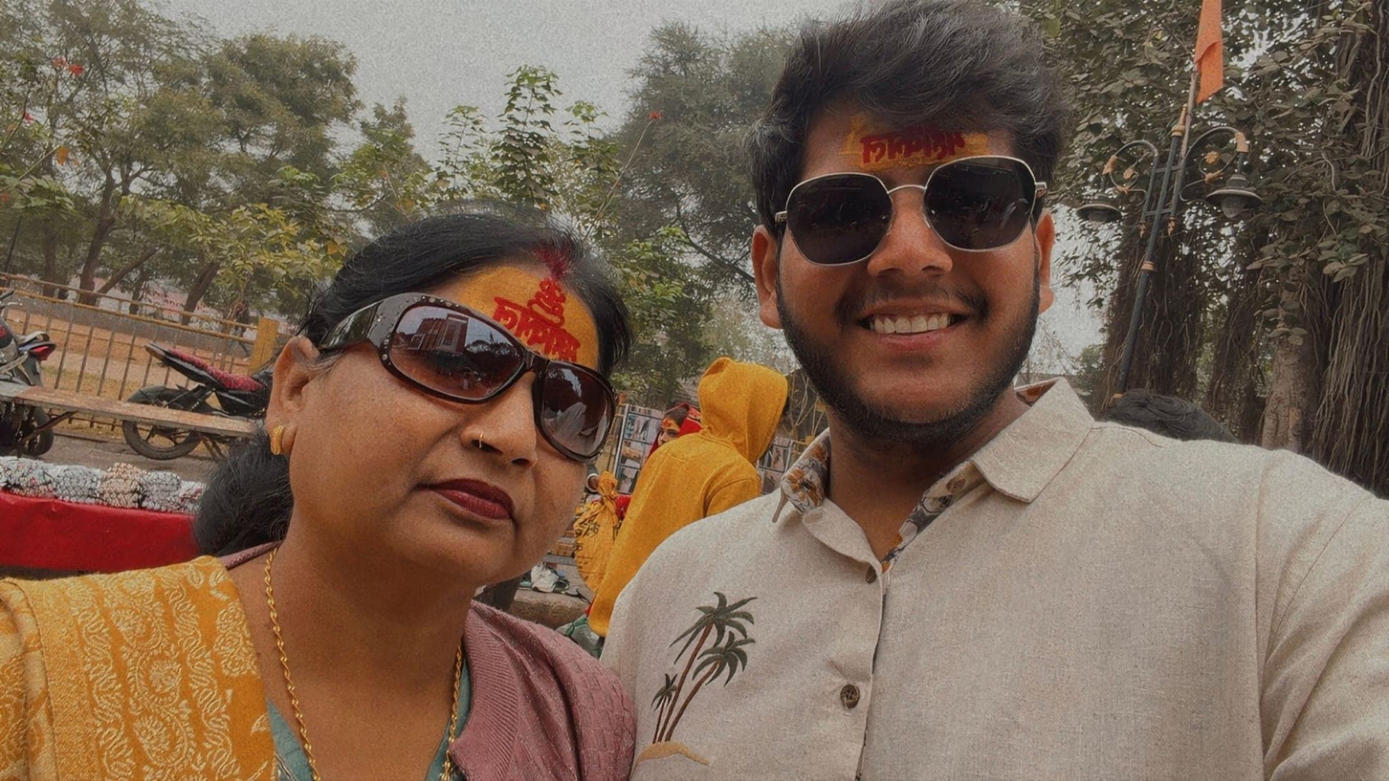 A selfie of a woman in sunglasses with a red tilak on her forehead and a young man in sunglasses and a cream shirt printed with palm trees, with a banyan tree and a market stall behind them