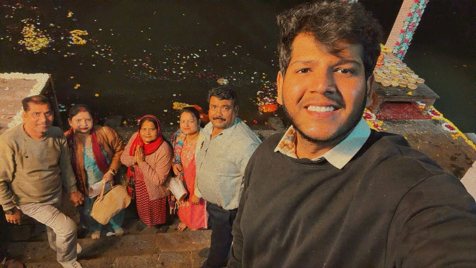 Six people sitting and standing on stone ghat steps at night, smiling for a selfie, with marigold flowers floating on the dark water behind them