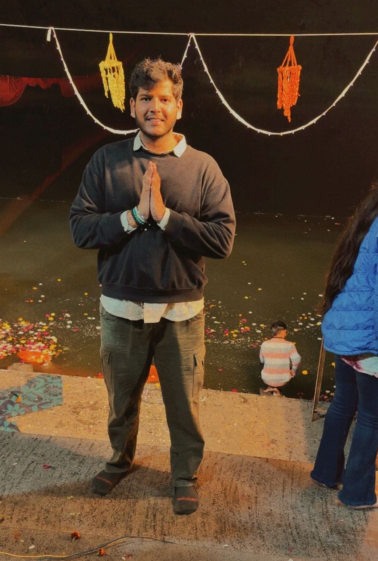 A young man in a dark sweater with his palms together on a ghat at night, with strings of lights and orange hangings above and flowers floating on the water behind