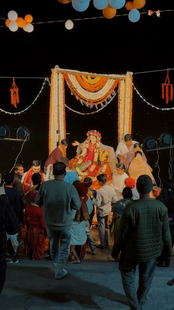 A goddess idol in red under an arch of marigold garlands at night, with people gathered around her and orange and blue balloons hanging above