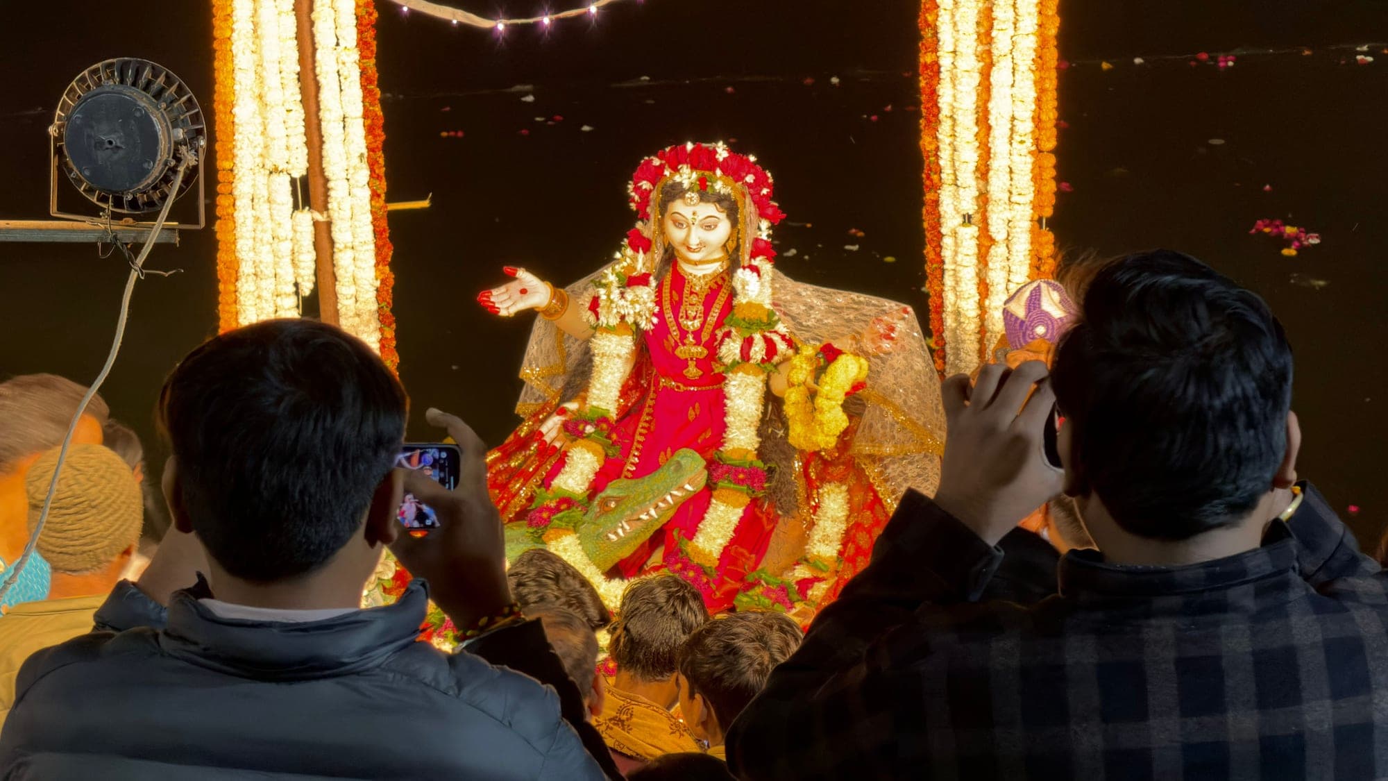 A goddess idol in red clothes and a crown, seated between tall columns of marigold garlands at night, with two people in the foreground raising their phones to photograph her