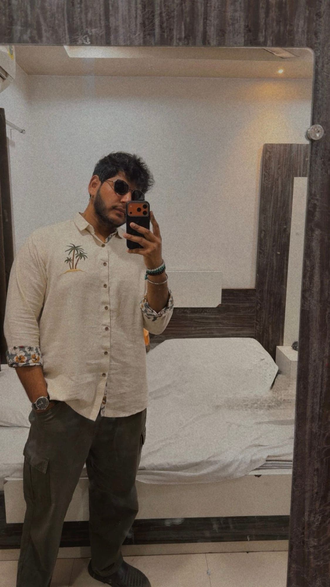 A young man in sunglasses and a cream shirt printed with palm trees taking a mirror selfie in a room with white bedding