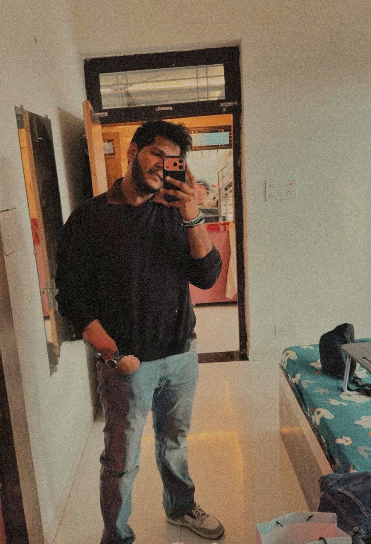 A young man in a dark sweater and jeans taking a mirror selfie in a bright room, with a bed with a flowered cover behind him