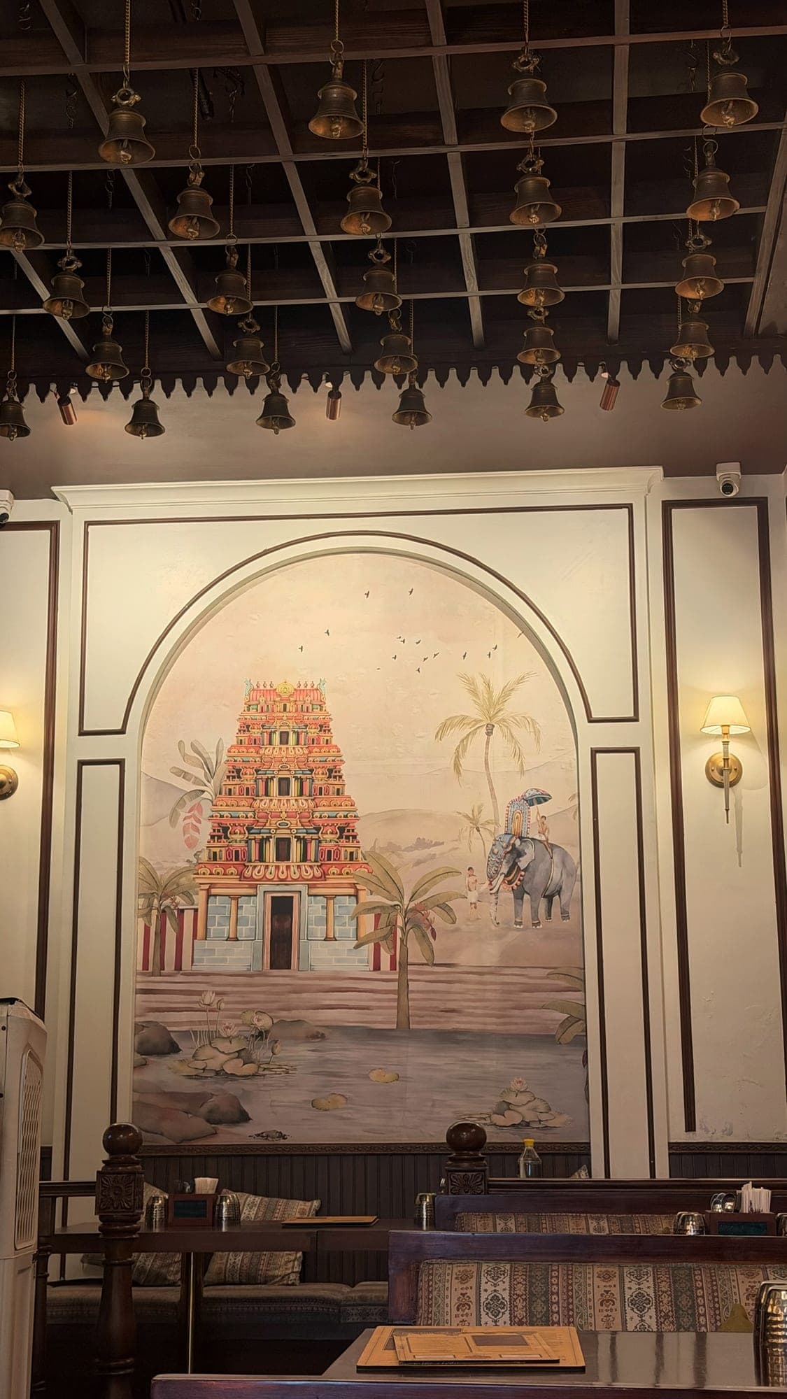 A tall arched wall mural of a colourful South Indian temple tower beside banana and coconut palms, a lotus pond in front and an elephant with a rider at the right, with brass bells hanging from a wooden grid ceiling above and booths below