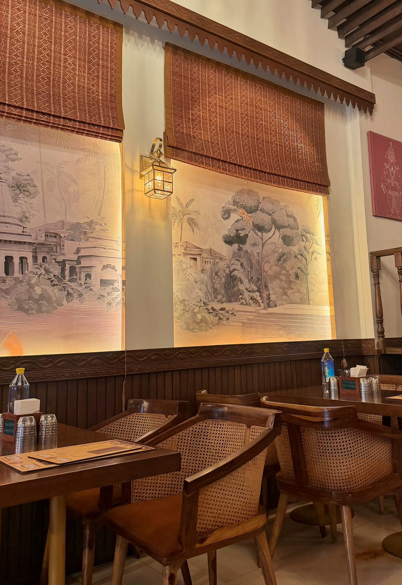 Two woven reddish blinds half drawn over backlit sepia murals of palms, riverside temple steps and a tree, with a brass lantern between them, and cane-backed wooden chairs and tables with menus below