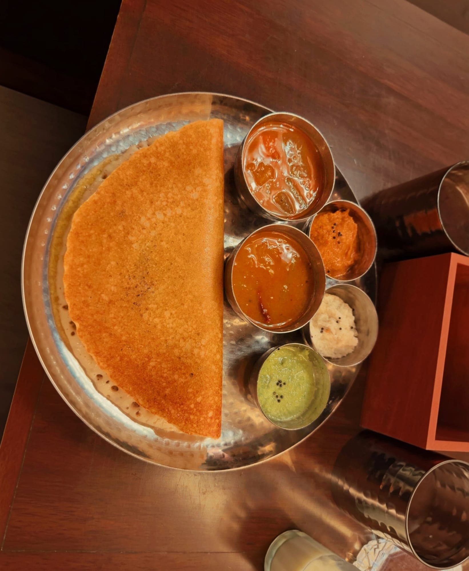 A crisp golden dosa folded in half on a round hammered steel plate, with small steel bowls of sambar and red, white and green chutneys beside it, seen from above on a dark wooden table