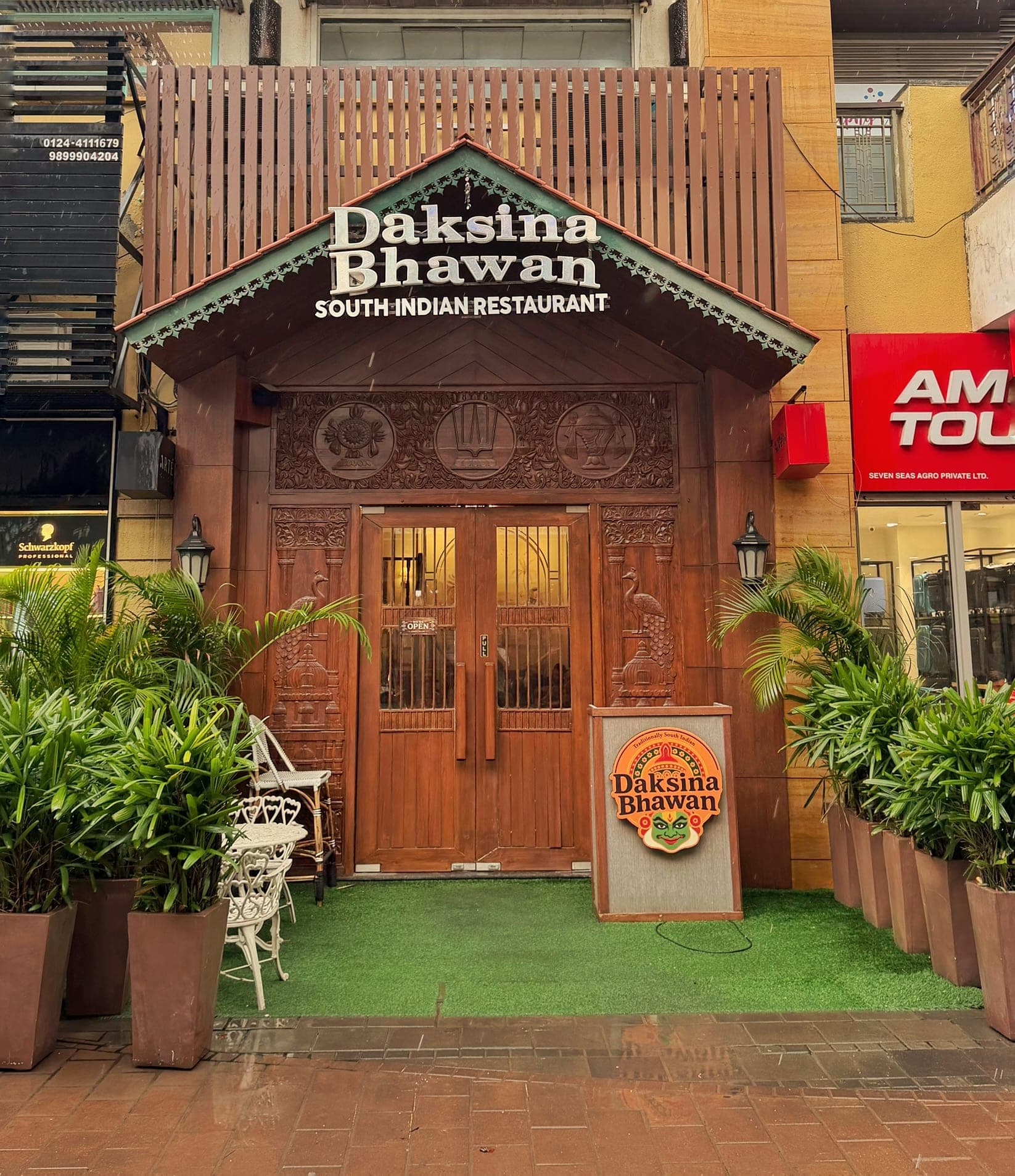 The front of Daksina Bhawan South Indian Restaurant: a gabled porch with a tiled roof and teal scalloped trim over the name sign, a carved wooden frieze with three round medallions above a pair of wooden doors with glowing glass panels, carved peacocks on the pillars either side, tall potted palms, a white wrought-iron chair and table on green turf, and a standing sign with a painted dancer's face, on a wet brick pavement