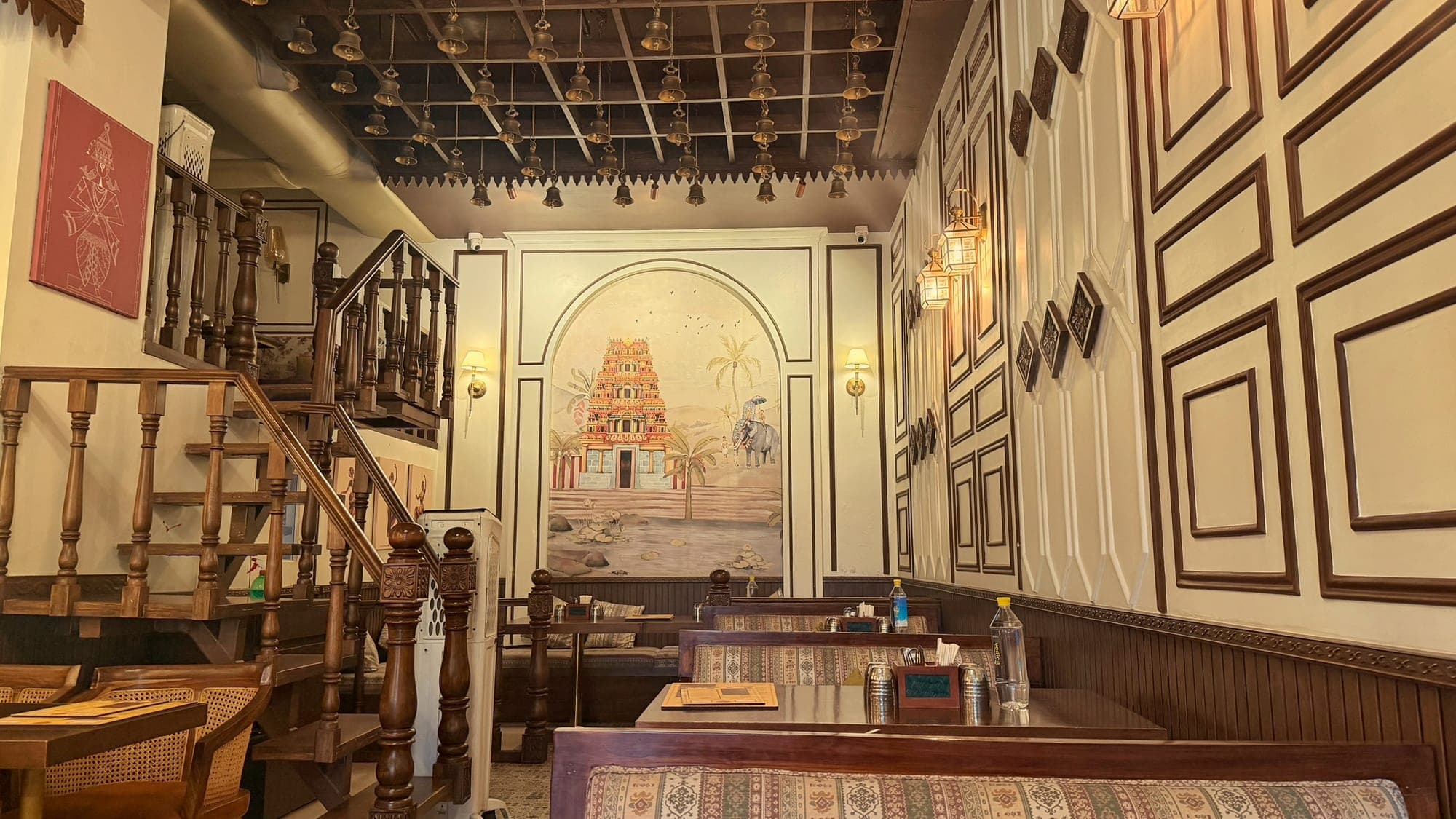 A narrow dining room with a dark wooden ceiling hung with rows of brass temple bells, a carved wooden staircase on the left, an arched mural of a South Indian temple tower with palms and an elephant at the far end, and cushioned booths along a panelled wall