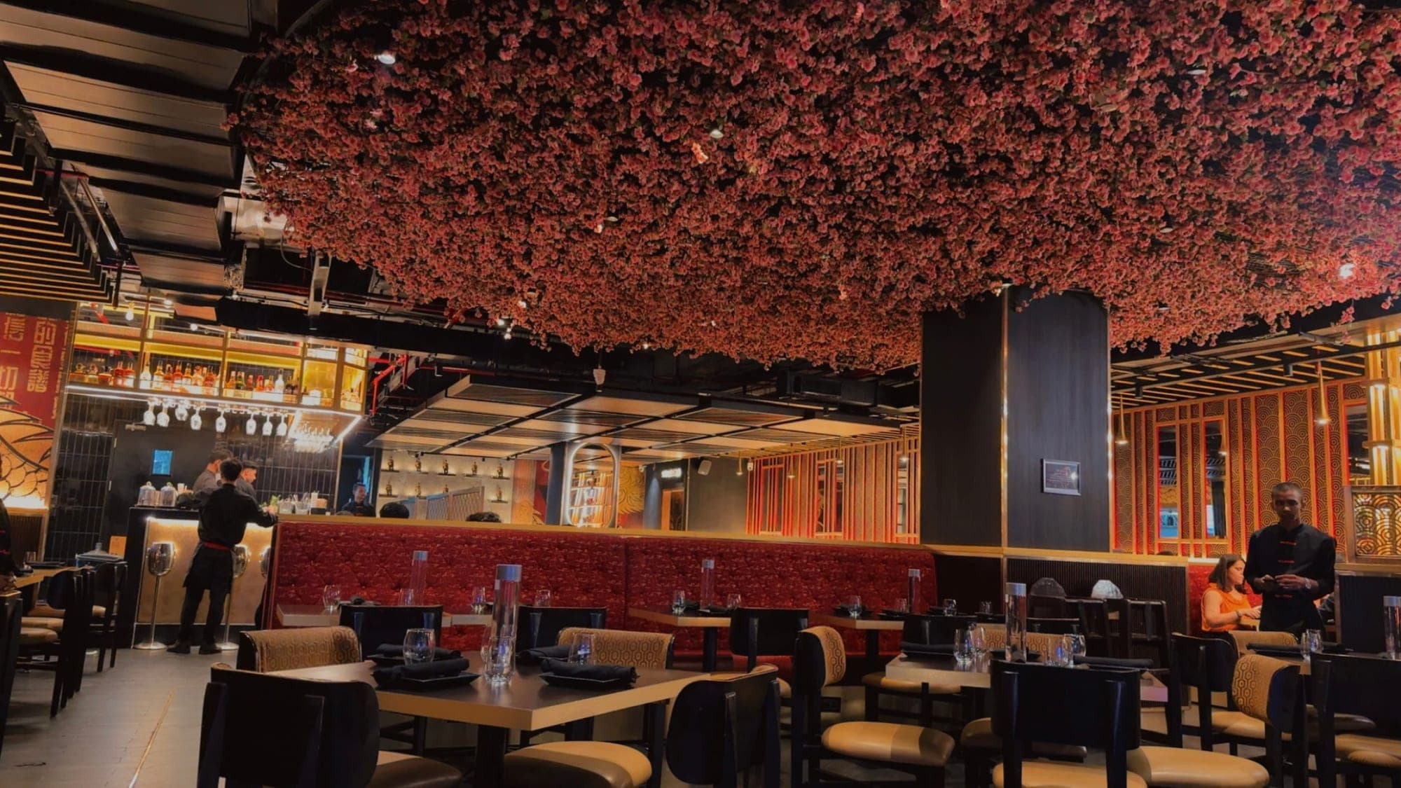 A restaurant dining room under a thick ceiling of pink blossom, with red leather booths, gold lattice screens and a lit bar at the back