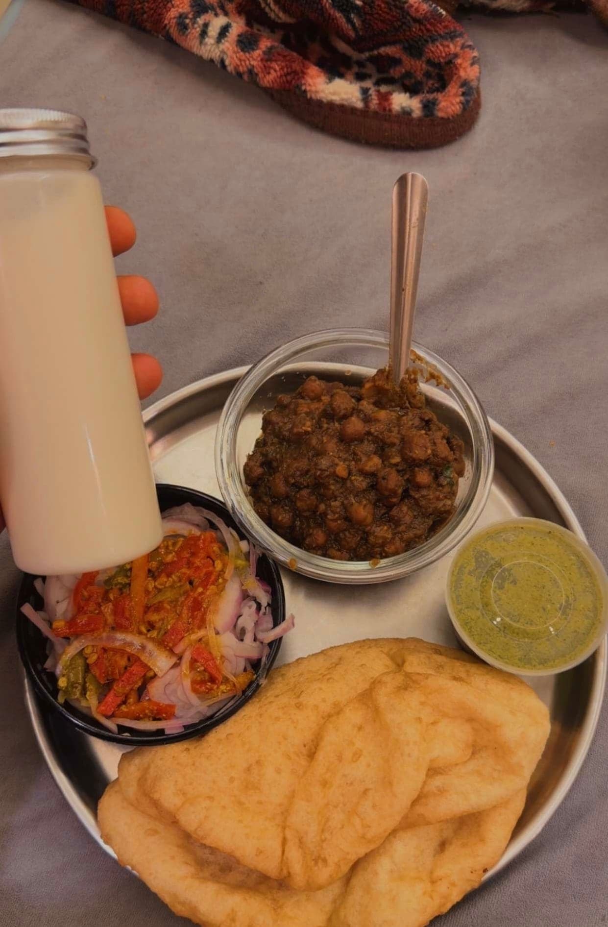 A round steel plate seen from above with two golden puffed bhature, a glass bowl of dark chole with a spoon, a black bowl of sliced onions and peppers, a small tub of green chutney, and a hand holding a tall bottle of sweet lassi, on a grey blanket