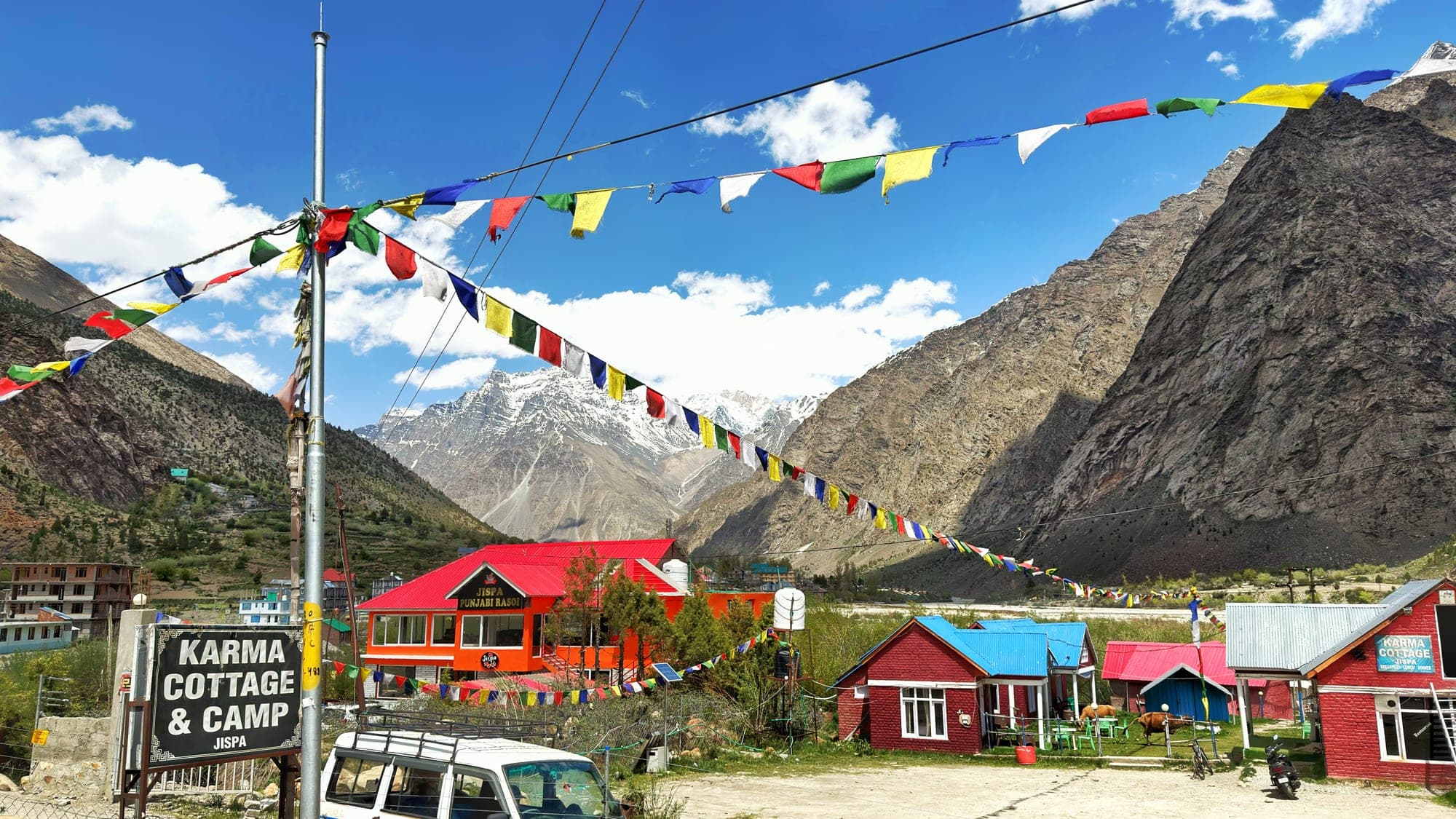 Lines of tattered prayer flags in blue, white, red, green and yellow crossing a bright blue sky above red-roofed cottages, an orange restaurant and a Karma Cottage & Camp, Jispa sign, with snow-topped peaks and a steep brown mountain behind