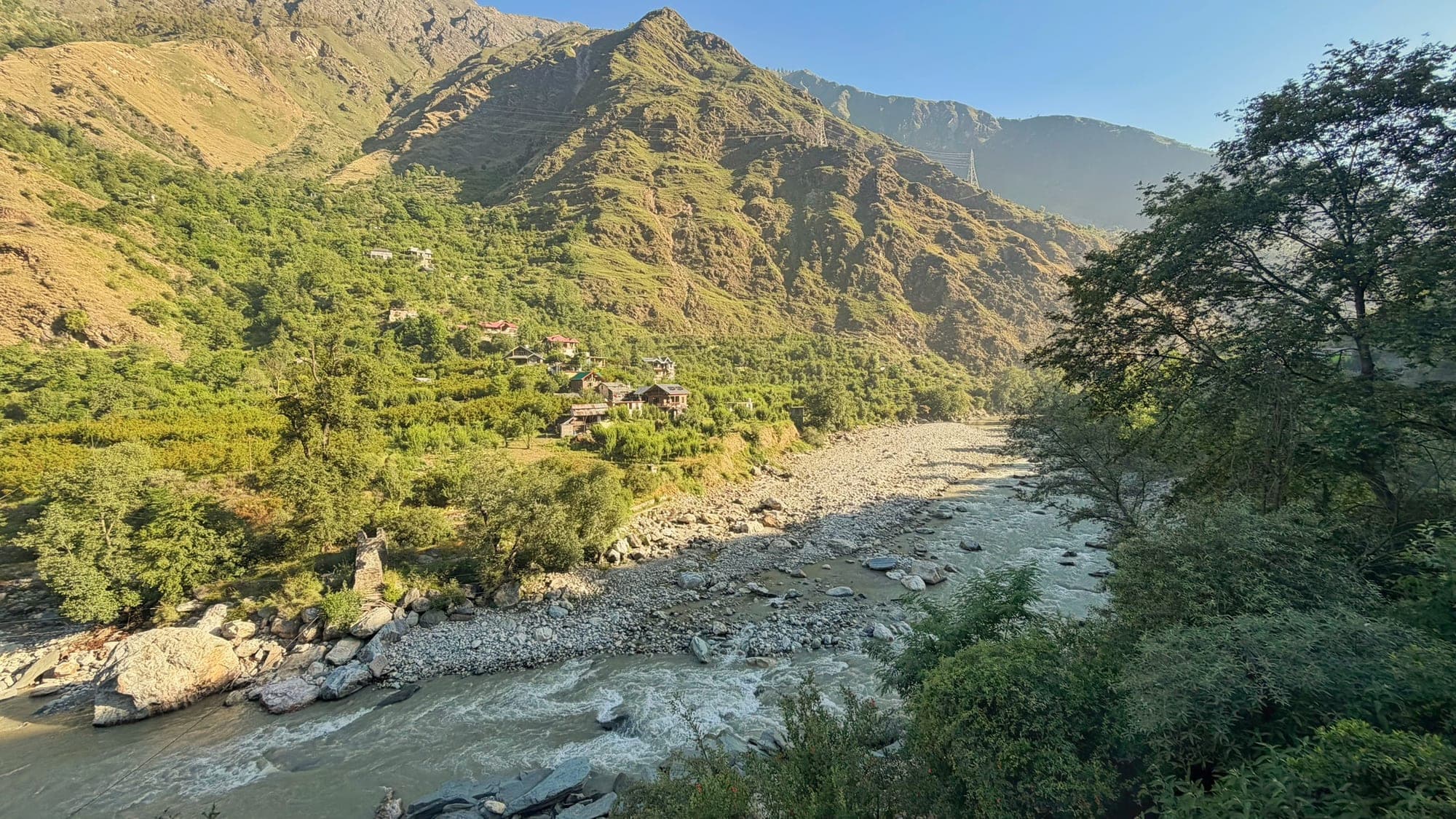 A wide view of the Parvati river rushing over a stony bed in a green valley, with a few wooden houses on the far hillside and steep, sunlit mountains behind, framed by trees on the right