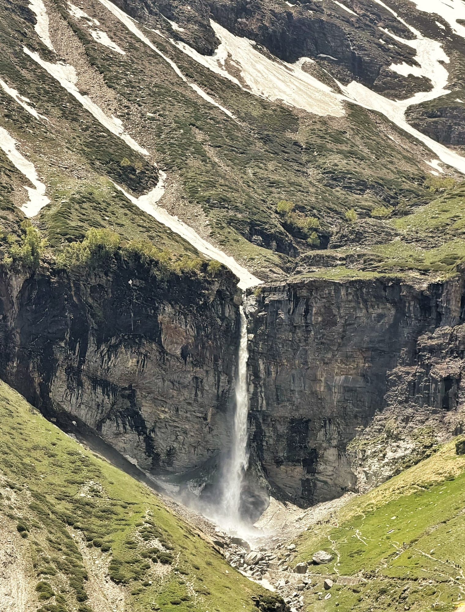 A thin white waterfall dropping down a dark cliff face into a spray-filled gorge, with tongues of snow and green scrub on the slopes above and bright green meadow either side of the base, where tiny figures stand